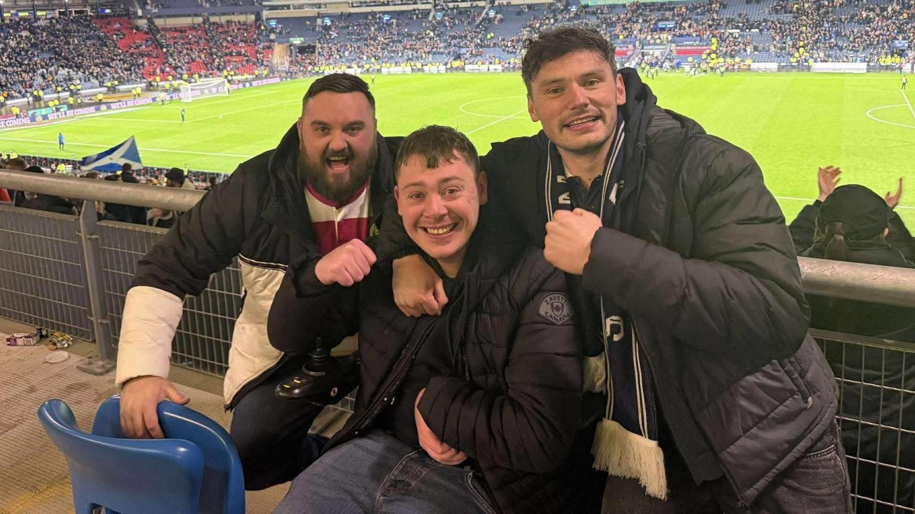 Stephen, who is sitting in a wheelchair, holds his right fist in the air in celebration after Scotland secured qualification for the World Cup. He is wearing a black jacket and blue jeans. He is flanked by two jubilant friends. The Hampden pitch and the red and blue stands can be seen in the background.