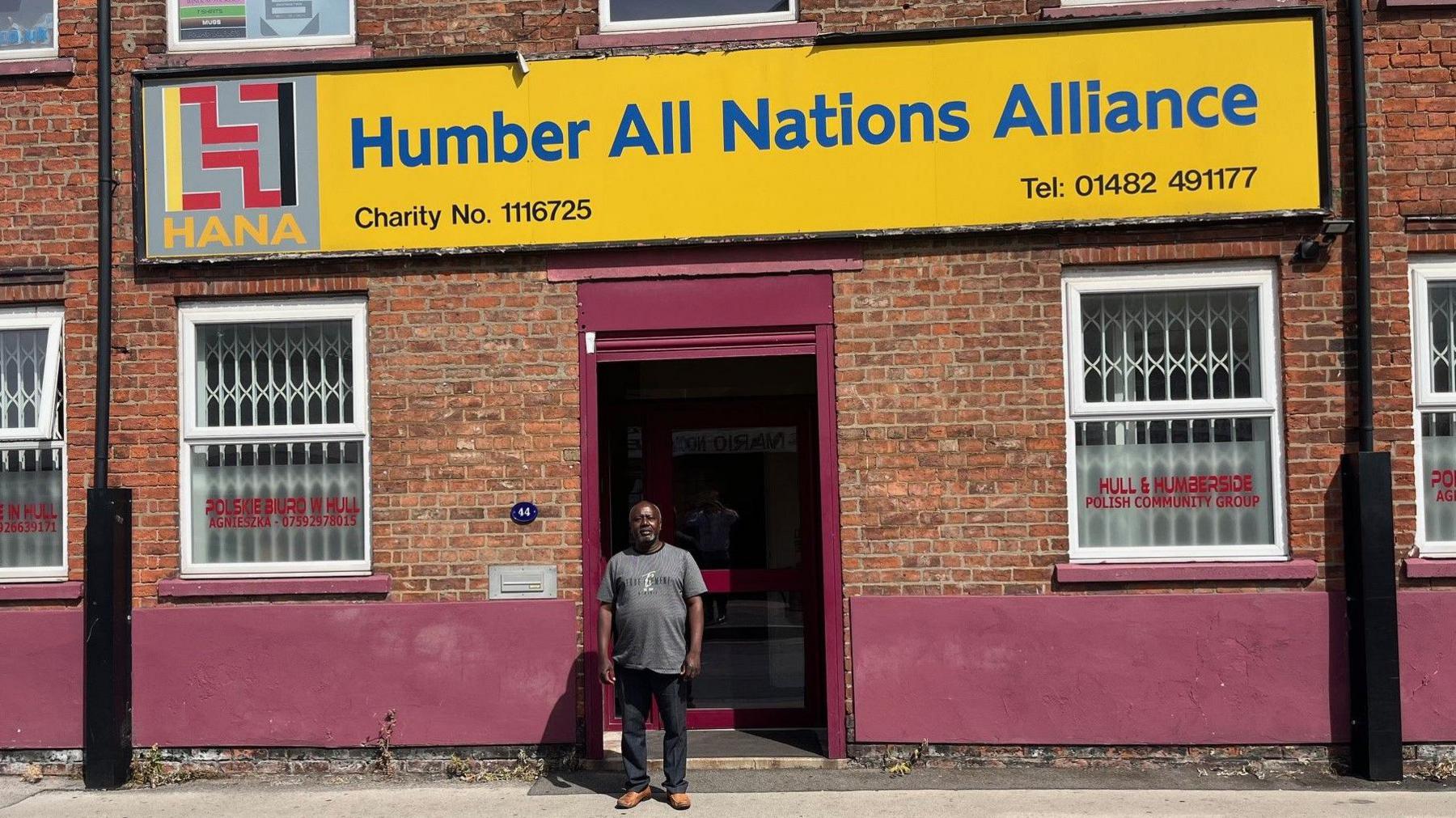 Francis Ahiakpa standing in front of Humber All Nations Alliance, a two-storey brick building with purple facias and a large yellow sign, on which is printed the name of the organisation in bold blue text. 