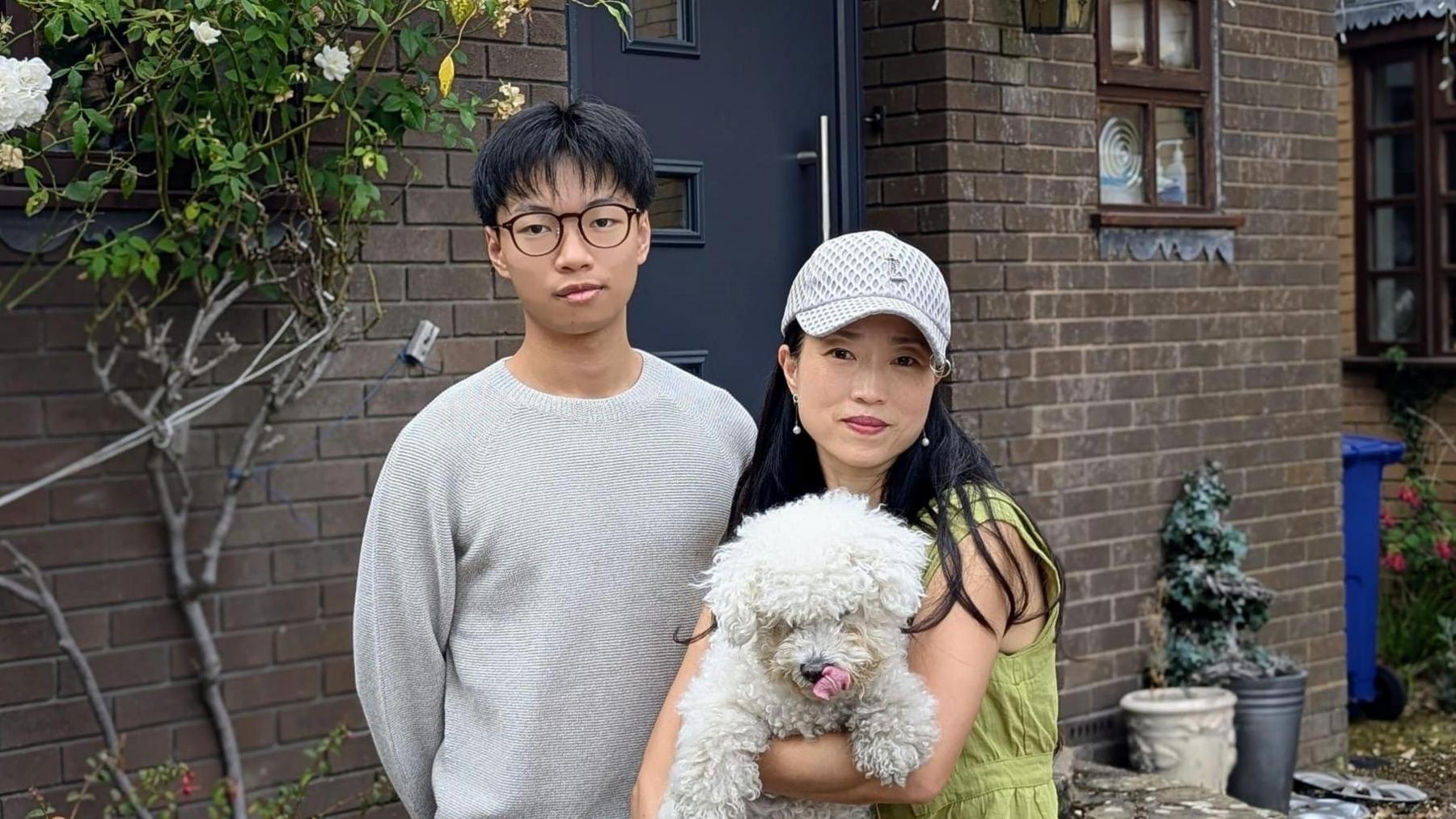 A man in a white jumper, black hair and round rimmed black glasses looks at the camera. Next to him is a woman, smaller than him, wearing a cap and holding a fluffy white dog. They are in front of their home. 