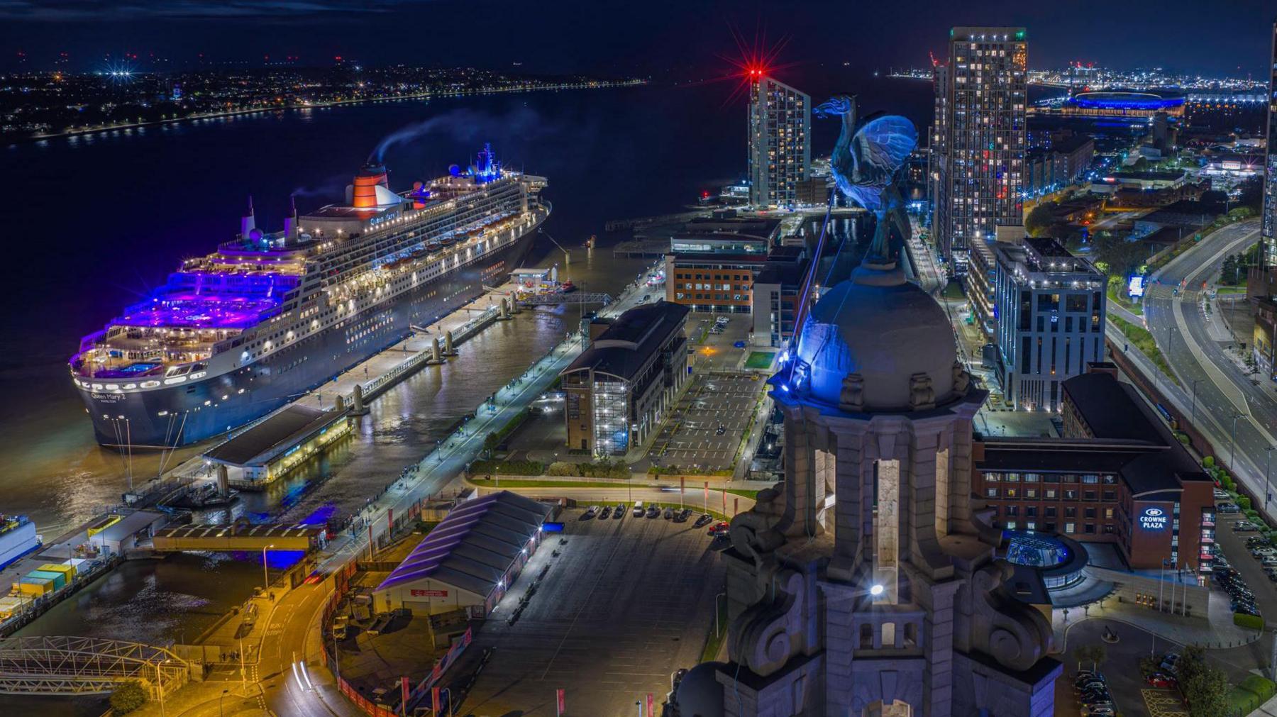 The Queen Mary 2 ocean liner on the River Merseys in Liverpool in front of the Liver Building. A Liverbird is lit up in blue and the Everton football stadium can be seen in the distance.