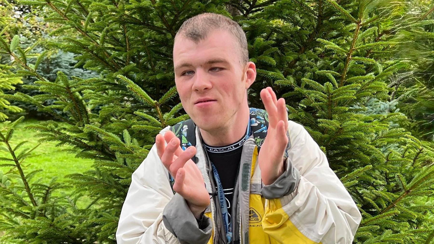 Billy is a young man with dark brown hair. He is wearing a cream jacket with a bit of yellow and is clapping. Behind him is a very green tree.