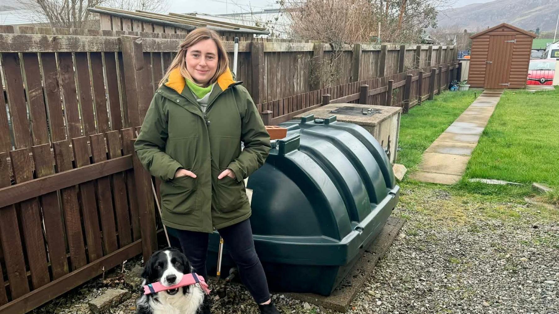 Allison NicIllEathain 's an cù aice air iall, nan seasamh ri taobh tanca ola aig an dachaigh ann an Ullapul