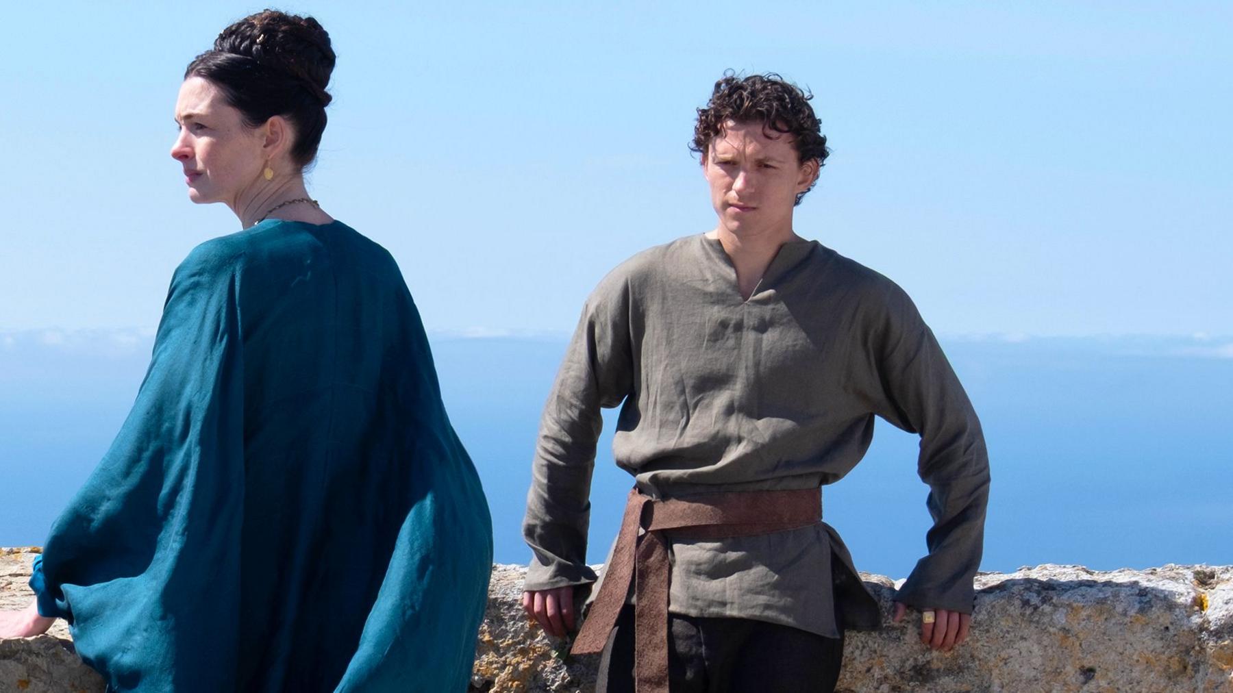 Tom Holland and Anne Hathaway in a film still from the film The Odyssey. Tom Holland is leaning against a wall with a scenic greek sea view  behind him. He is wearing a dark olive green shirt and dark trousers. Anne hathaway is looking to the left and wearing a flowing flowing blue dress