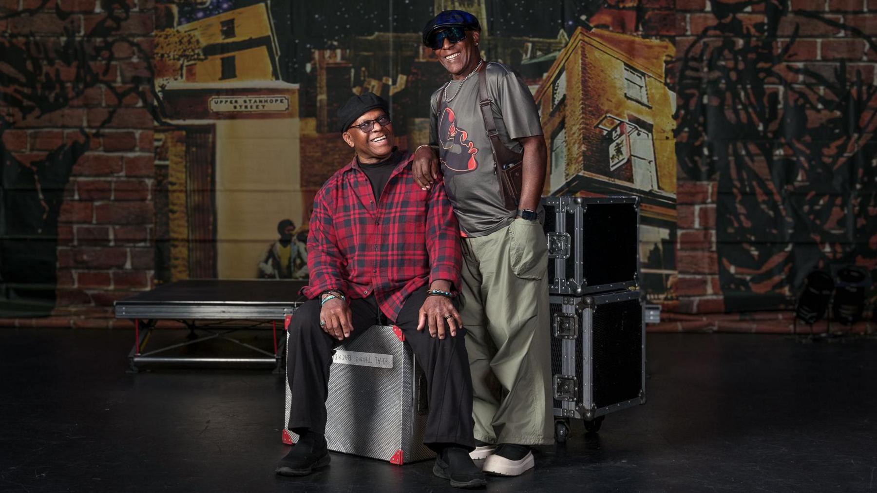Two members of The Real Thing. One is sat on a metal box and is wearing black trousers and a red and black checked shirt, a black hat and glasses. The other is stood up wearing green trousers, a grey t-shirt and a black hat and dark sunglasses.