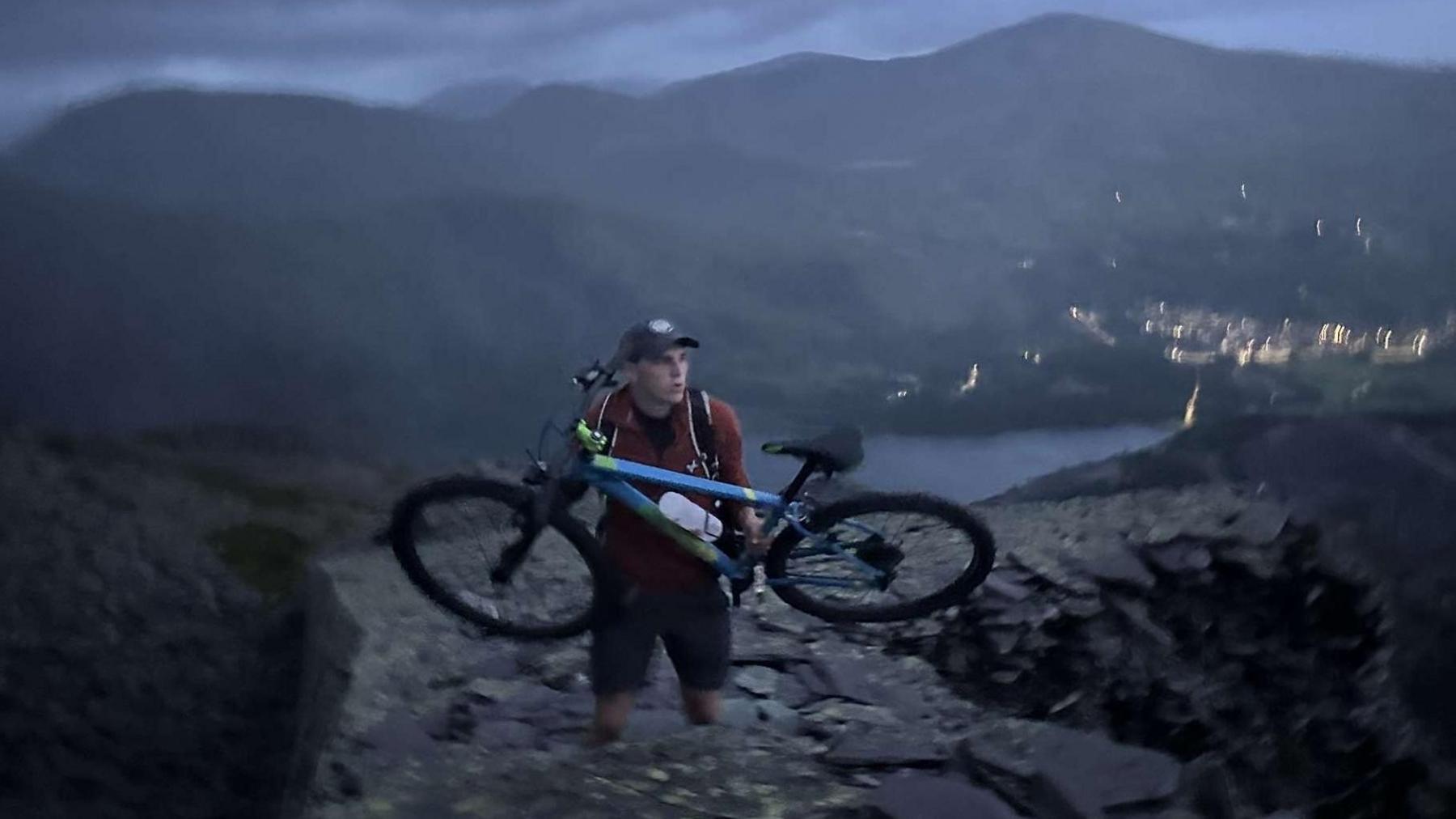 Aran in dramatic night photo. He is holding a mountain bike in front of him while walking up a stone path on a mountainside over looking a lake and a town a night, shown by the lights. Aran is a bit blurry suggesting motion and wearing a ball cap a maroon hoodie with the hood down and zip down slightly and dark shorts