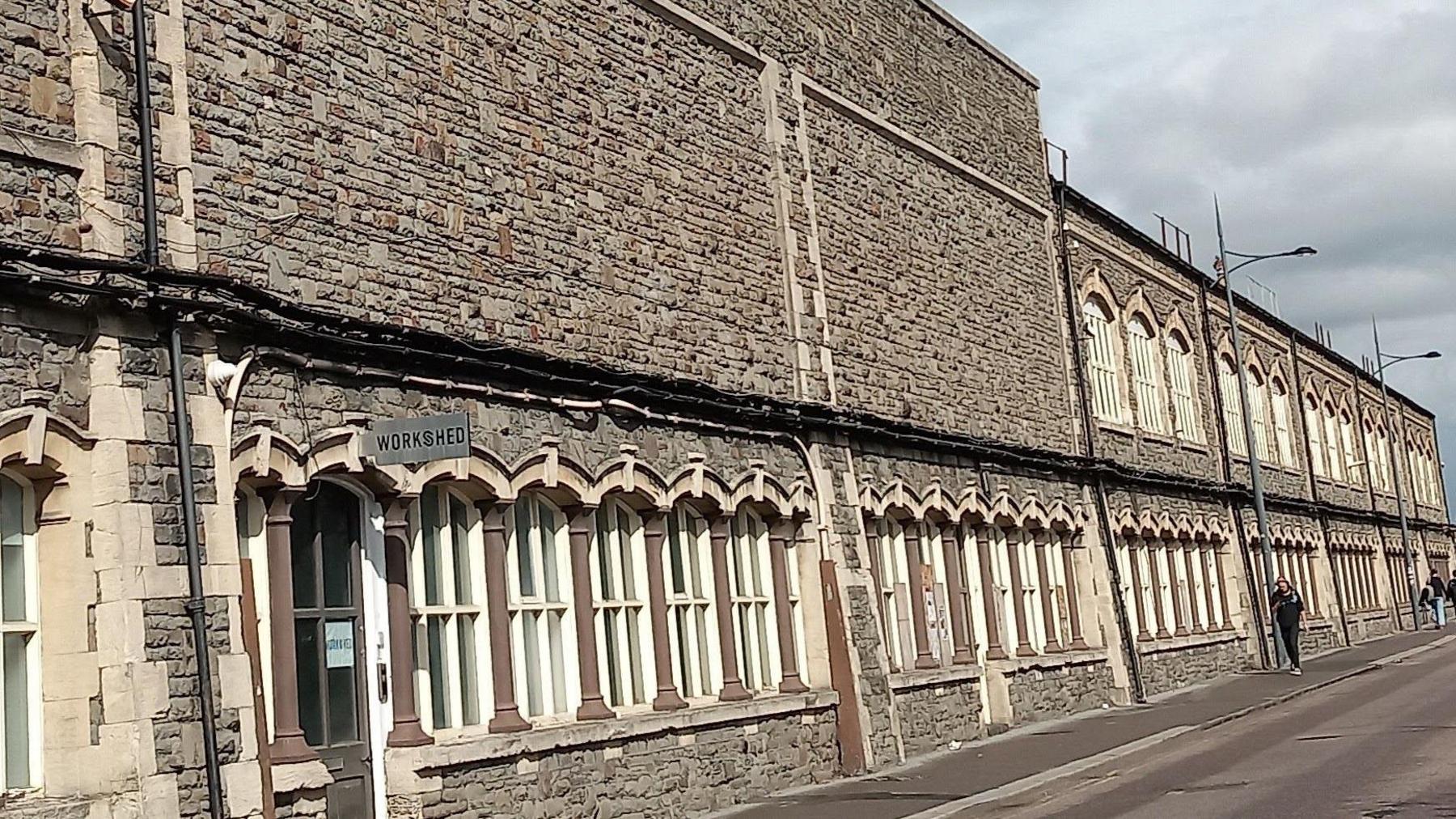 An image of a tall brown and grey stone wall, the bottom section of which includes a row of arched windows and a brown door. A sign over the door reads "workshed".