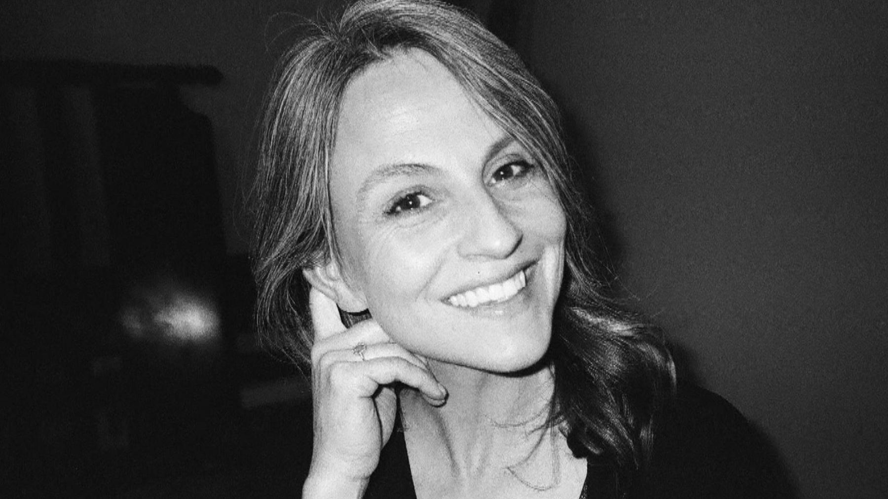Katie Bastion-Strong, smiles in a black and white image, looking into the camera while her hand is held behind her ear.