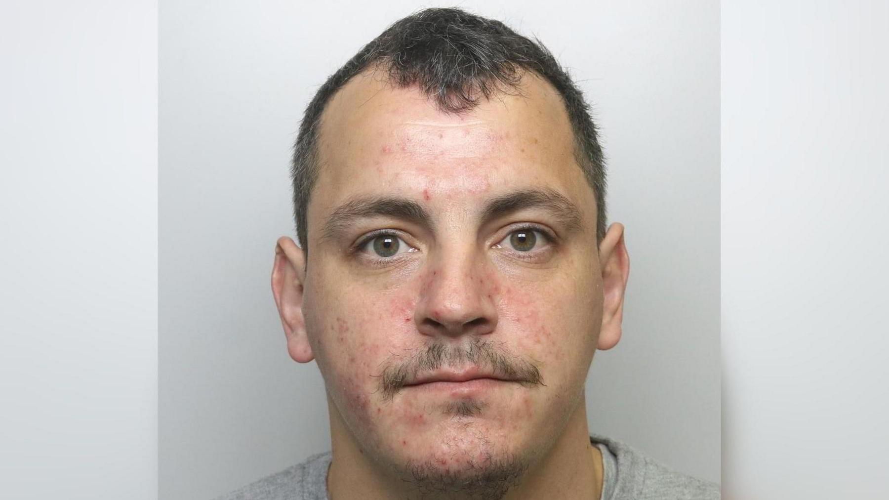 A police headshot of a man with short dark hair wearing a grey T shirt.