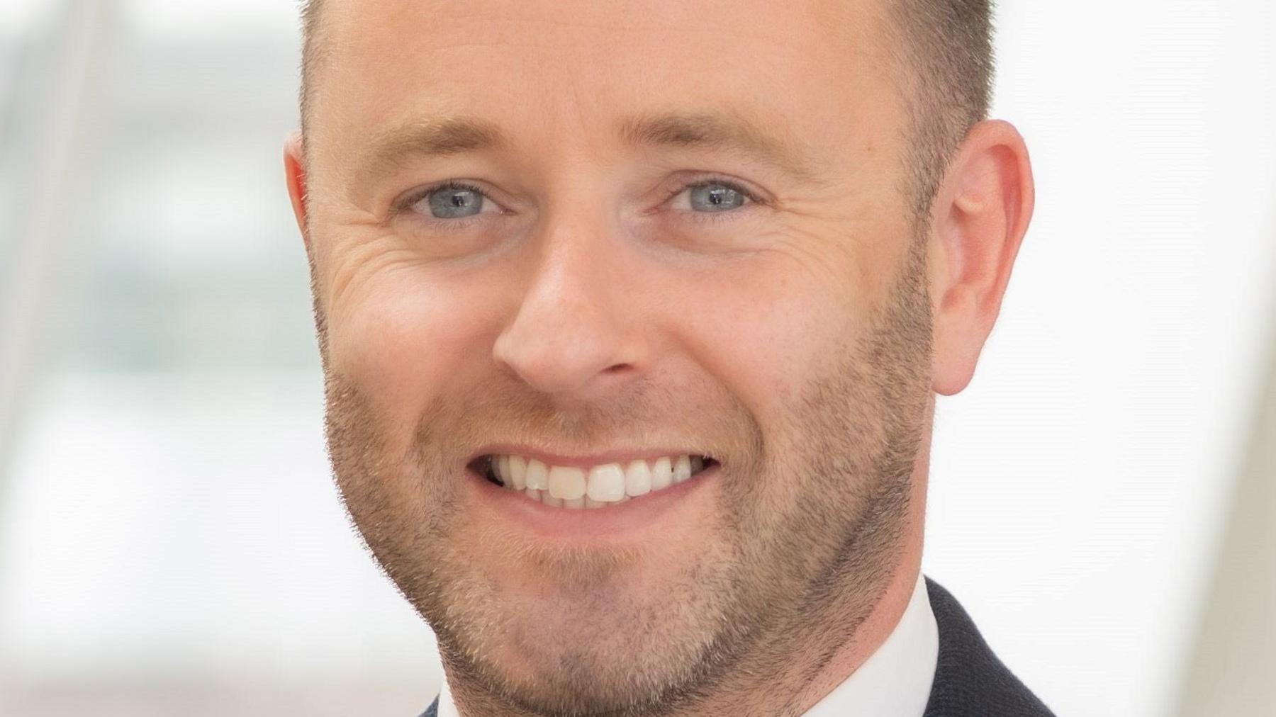 A man with a short stubble and wearing a black suit and white shirt smiling into the camera.