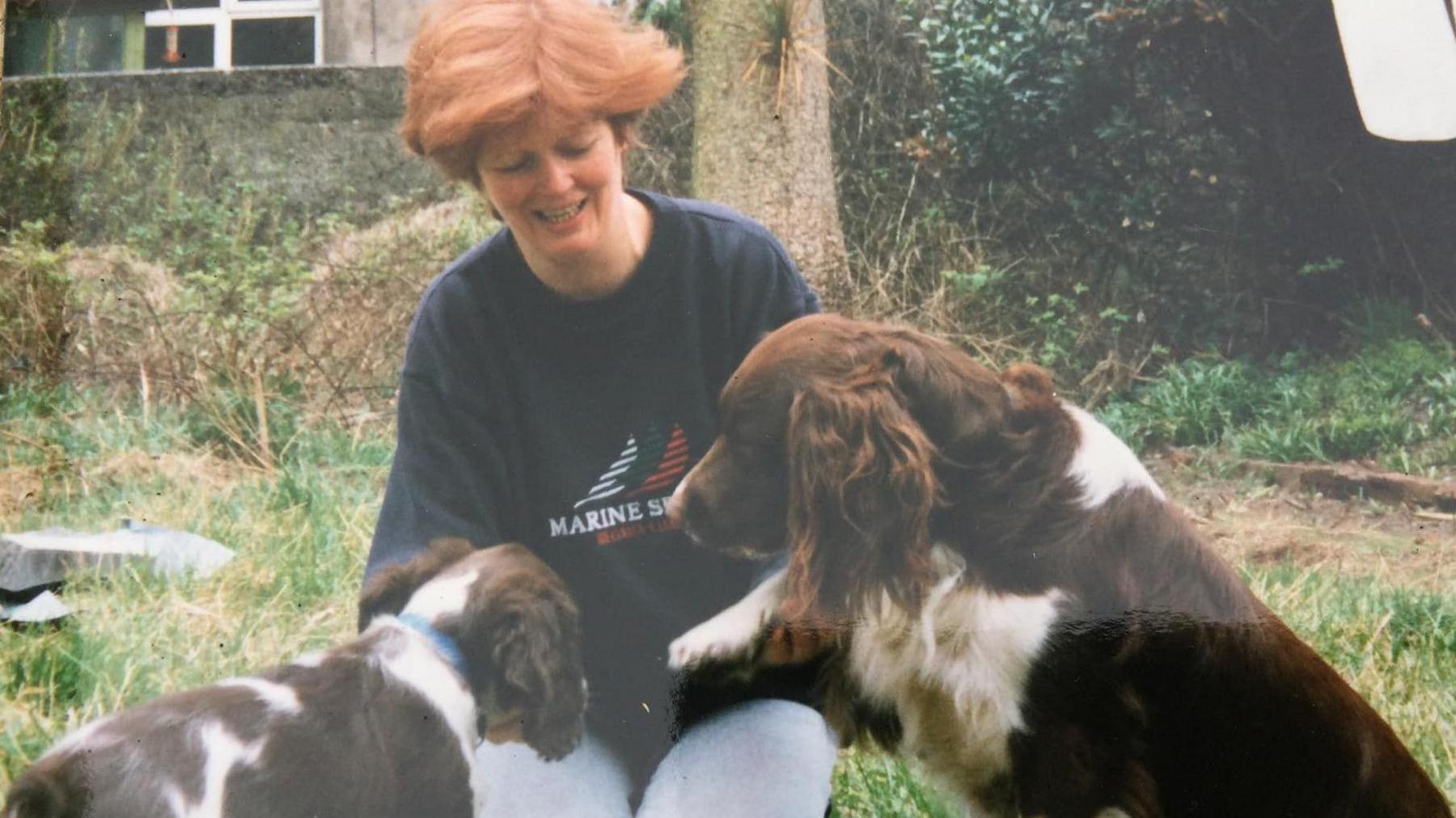 Shona Campbell in her 30s with short ginger hair as she kneels on the grass playing with two brown and white springer spaniels.