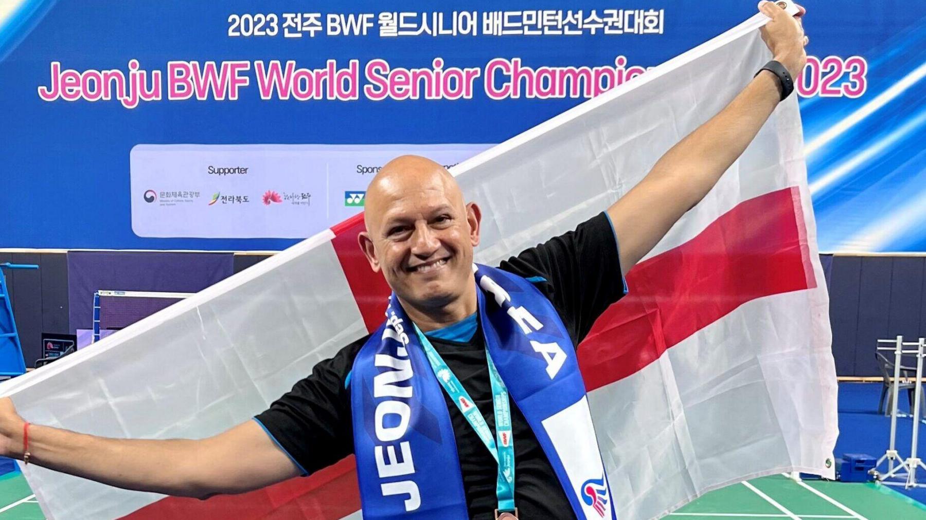 Rajeev is smiling looking directly at the camera. He is wearing his medal and is dressed in a black t shirt holding an England Saint George's cross flag behind him