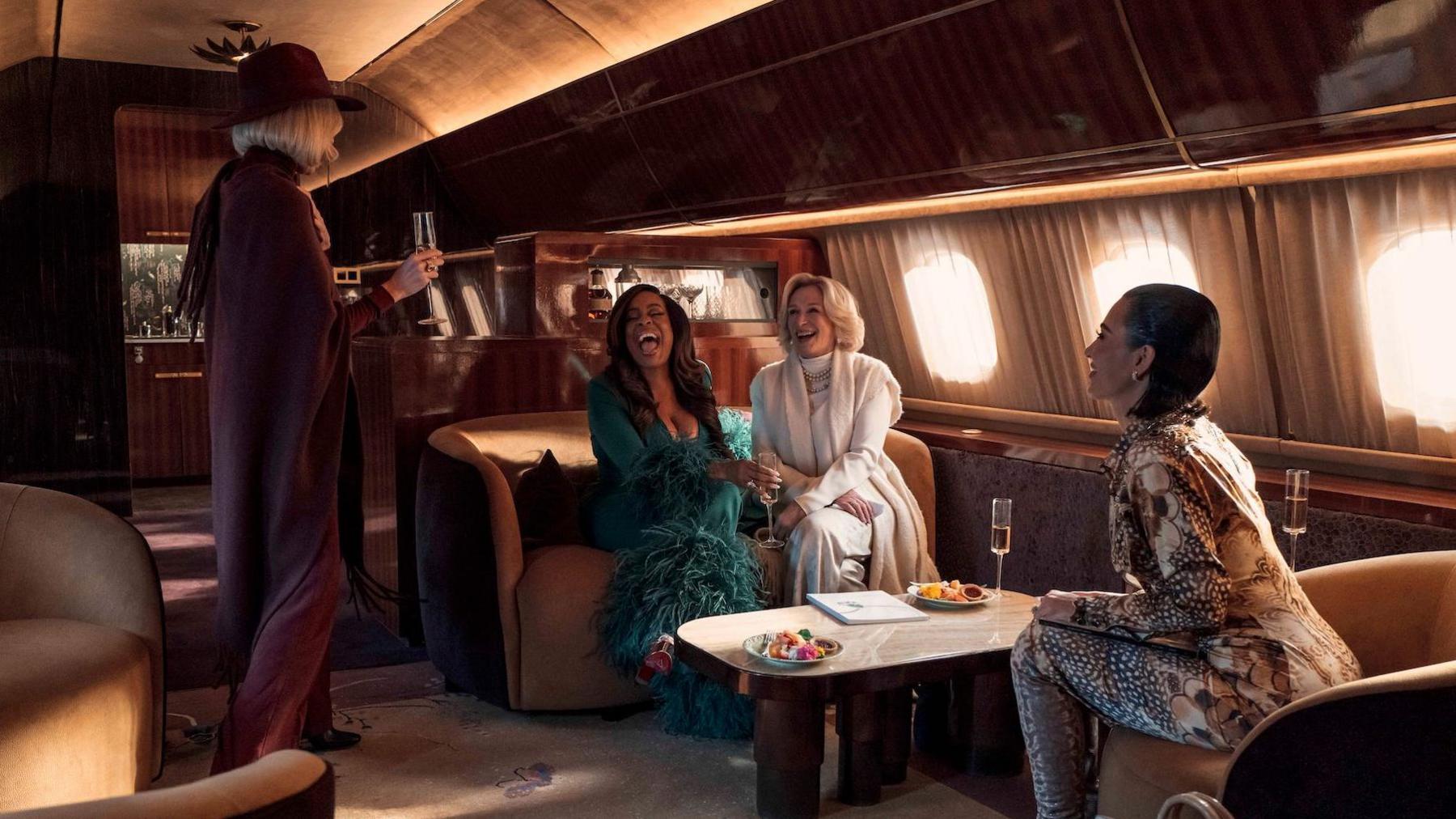 Naomi Watts, Niecy Nash, Glenn Close and Kim Kardashian pictured in new Disney+ show. They are all sitting on a private plane, drinking champagne.