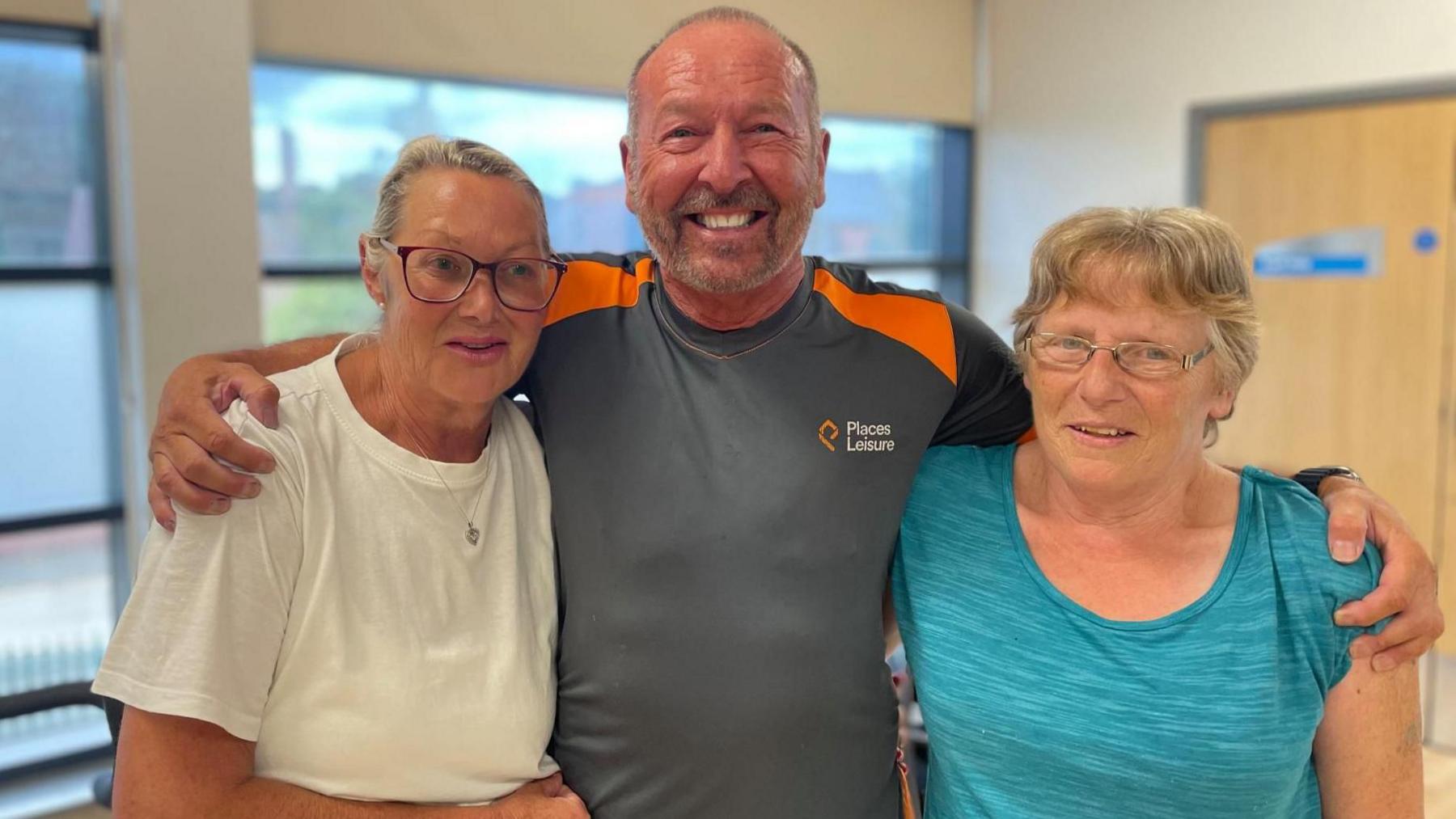 A man in a grey sports top hugs two nervous looking women both in sports tops and wearing glasses