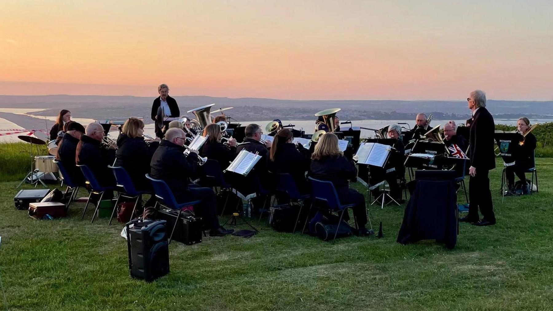 A brass band playing on top of the hill at Portland overlooking Chesil Beach as the sunset turns the sky orange