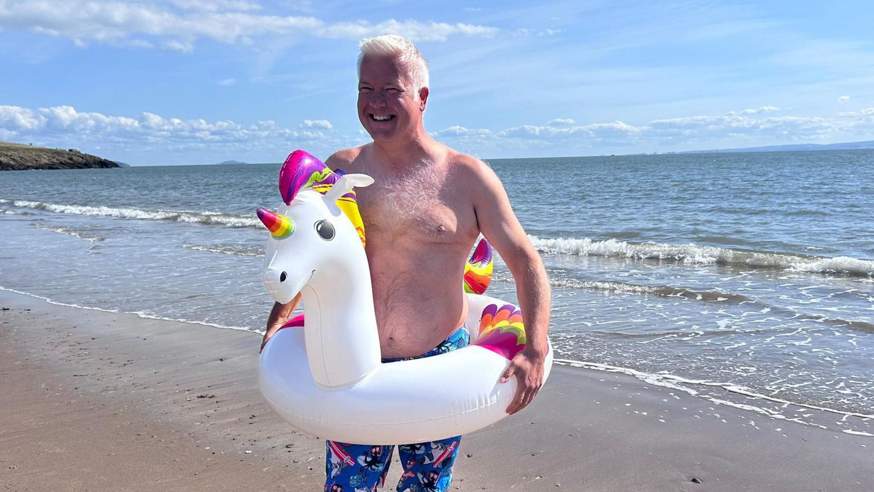 Derek on Barry beach in swimming shorts with a a white unicorn rubber ring around his waist. 