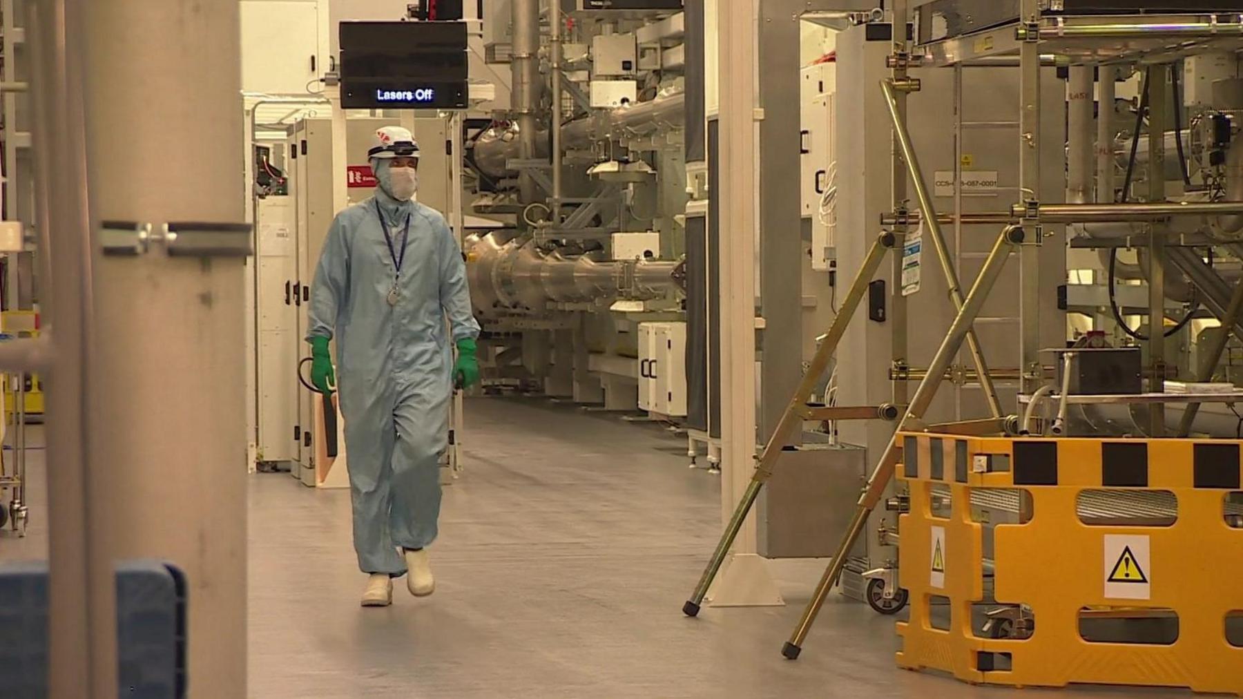 A masked engineer in a light blue hazmat suit walking through a room of industrial equipment