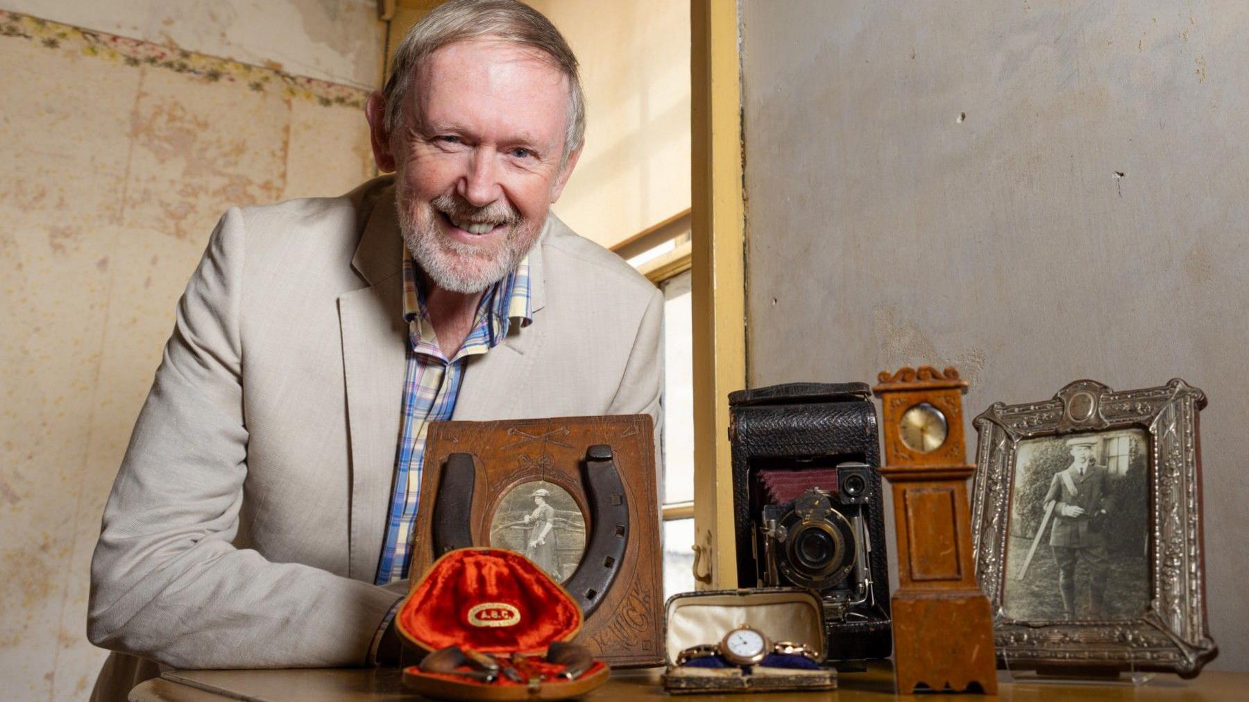 A man with grey hair wears a beige coloured jacked with a checked blue, white and yellow shirt beneath. he has a moustache and short cut beard. He's in a room with pale walls and a window with a half open blind behind him, letting in daylight. On a desk in front of him are a number of items, including a wristwatch, an antique looking camera, photo in a frame and a horseshoe standing upright on a wooden backing board
