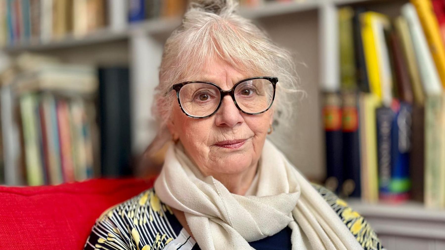 A woman aged 75 with white hair tied back, wih a fringe and large black coloured spectacles. She wears a cream scarf and a yellow and black patterned top. Bookshelves are visible behind her, she sits on a red sofa.