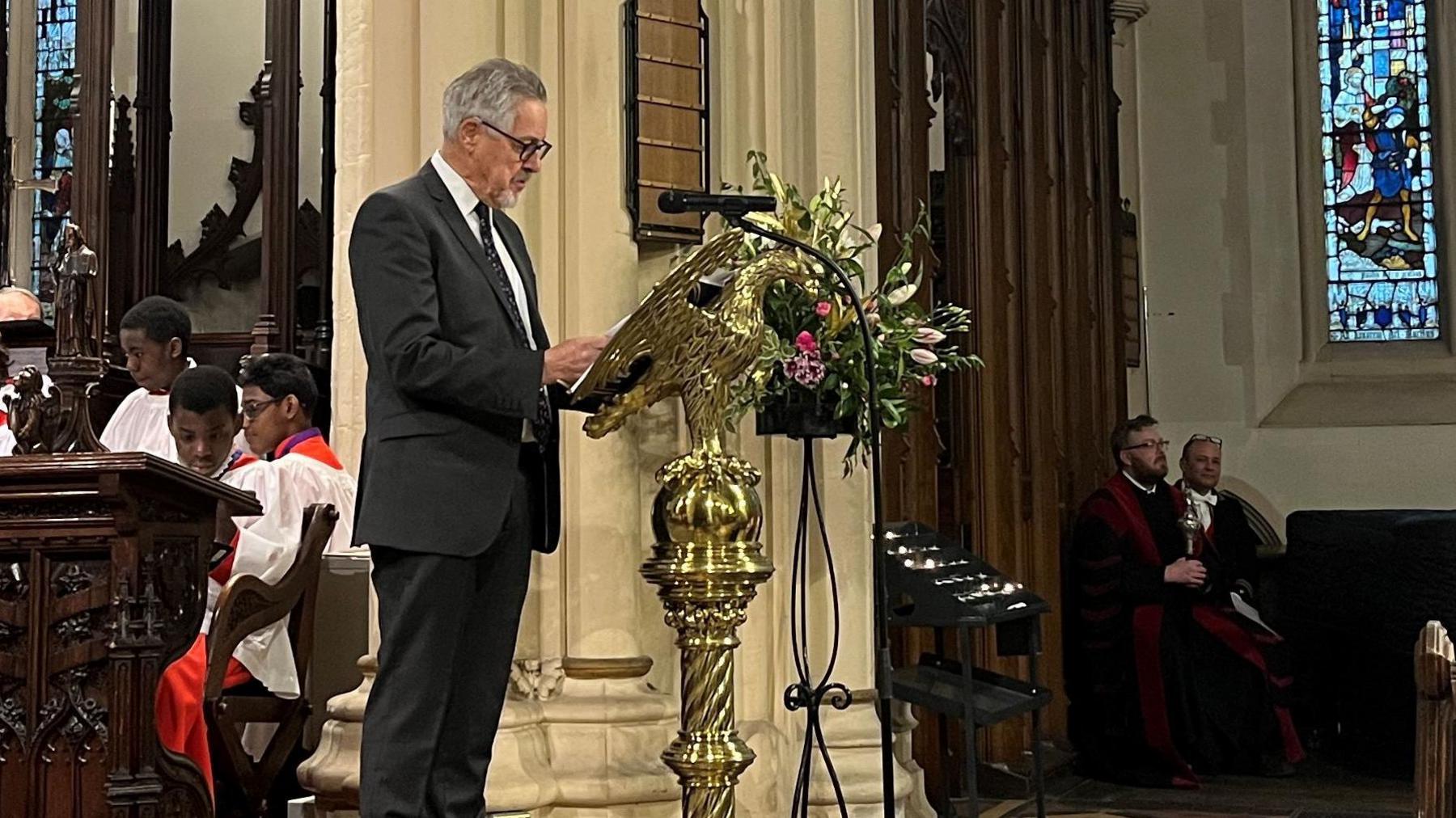 Griff Rhys Jones stands reading at a church lectern. He is wearing a dark-coloured suit and patterned tie.