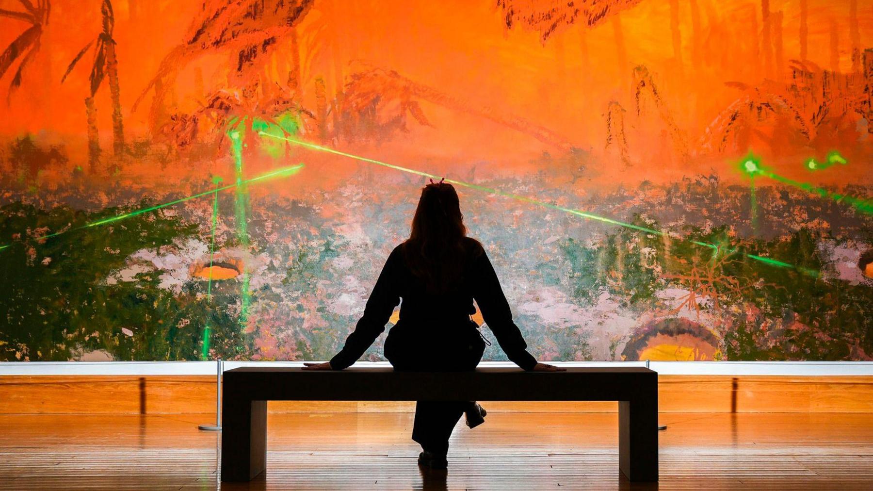 A silhouetted woman sitting on a gallery bench in front of Mohammed Sami's large painting