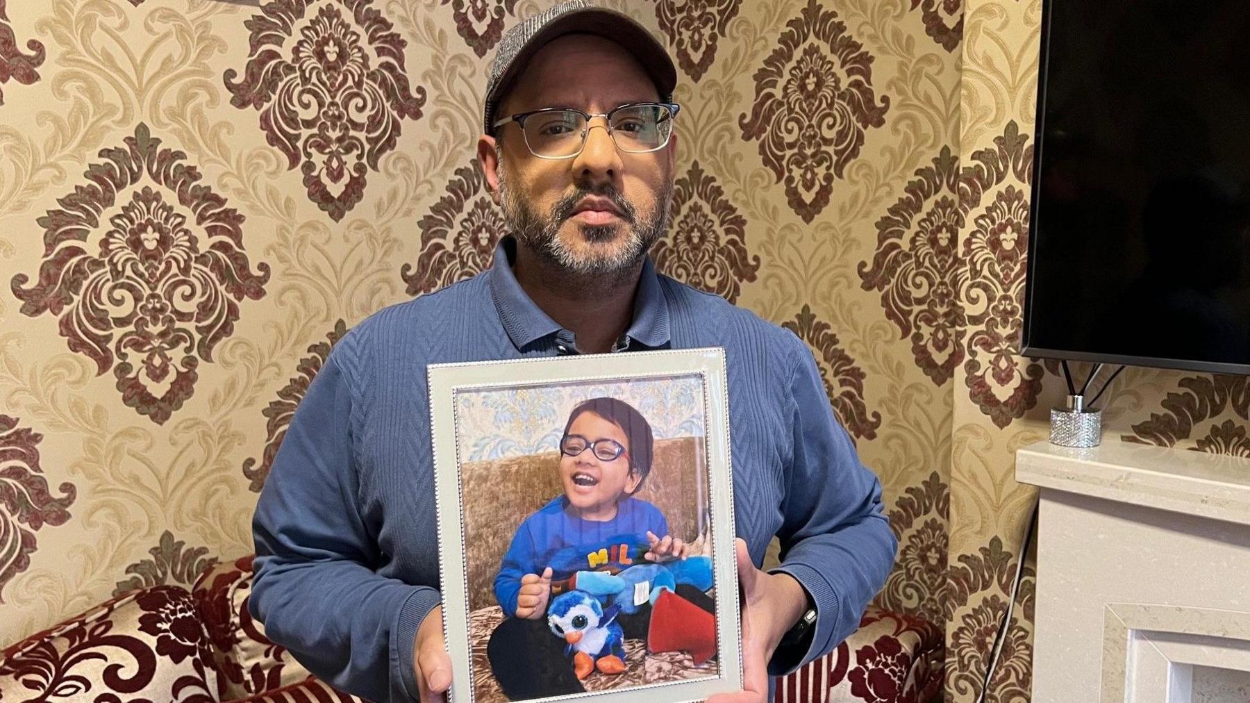 Haroon Rashid, wearing a lavender coloured shirt, a cap and glasses, holds a framed photo of his son Muhammad Ayaan Haroon, who is smiling and laughing as he plays with a blue cuddly toy.