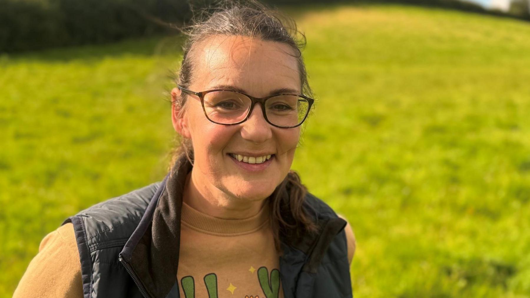 a woman in a jumper, gilet and glasses smiles at the camera. She is standing in front of a grassy hill and the sun is shining.