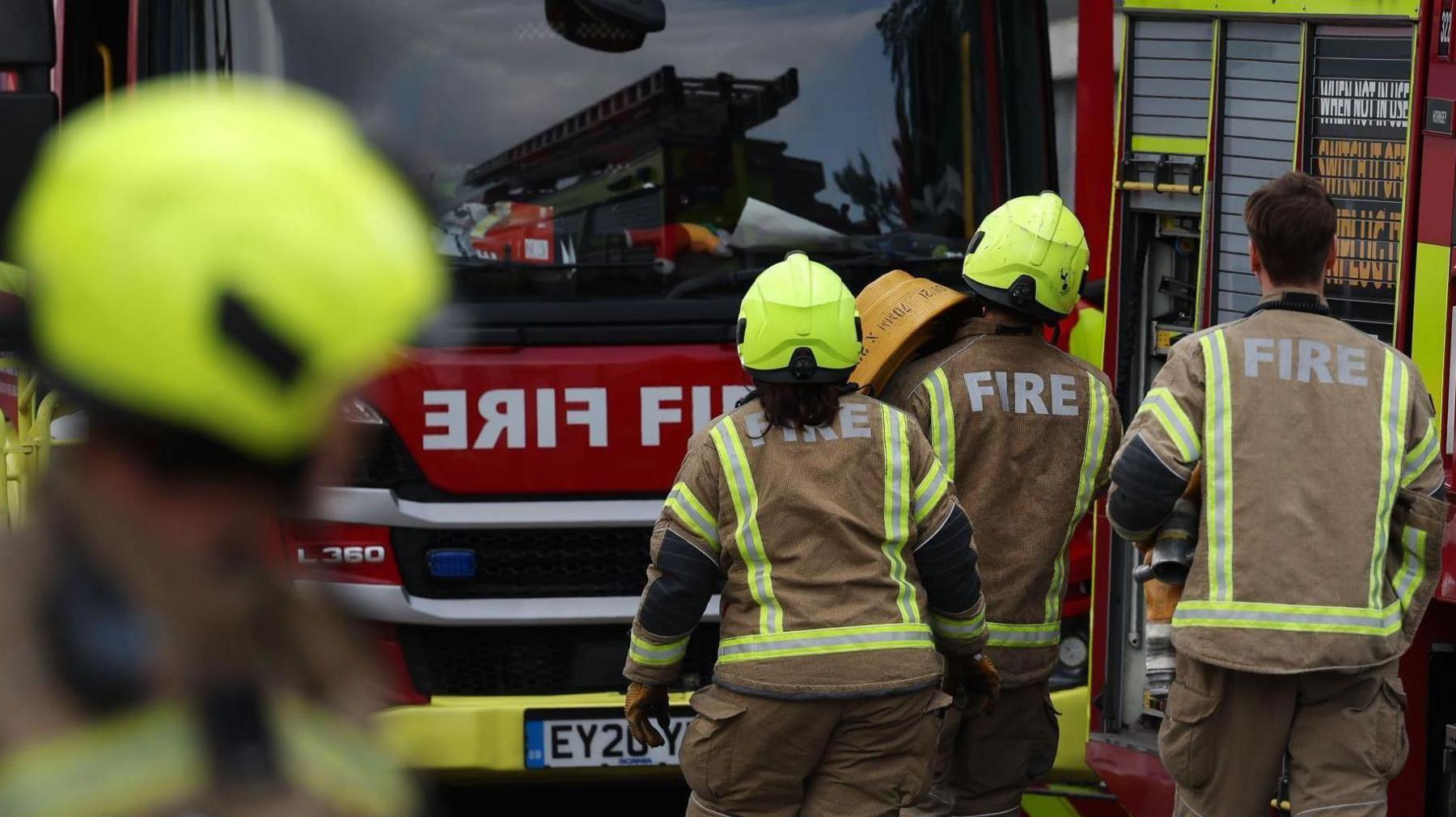 Three firefighters walk towards a fire engine with another crew member blurred in the foreground. They wear brown uniforms with fluorescent strips, with 'fire' printed on the uniforms in big white letters.