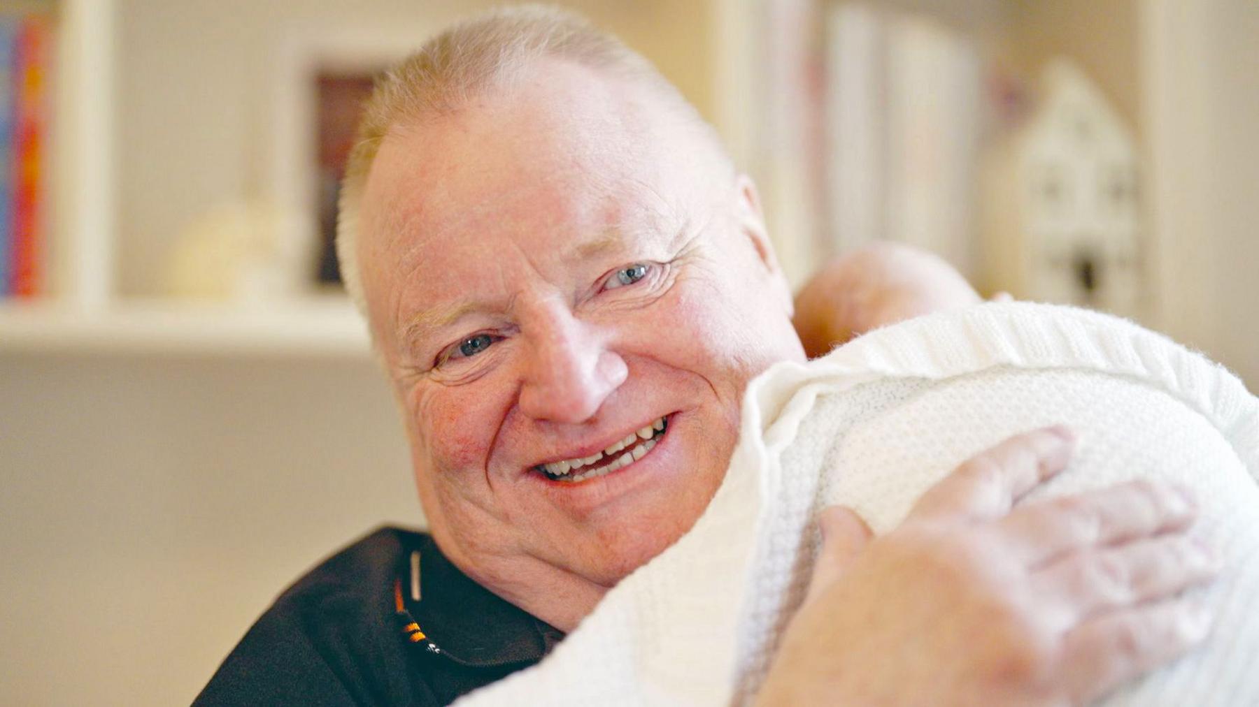 Ray, a 62-year-old man with closely cropped hair, smiling broadly as he holds his infant granddaughter Willow against his shoulder, patting the blanket she is wrapped in.