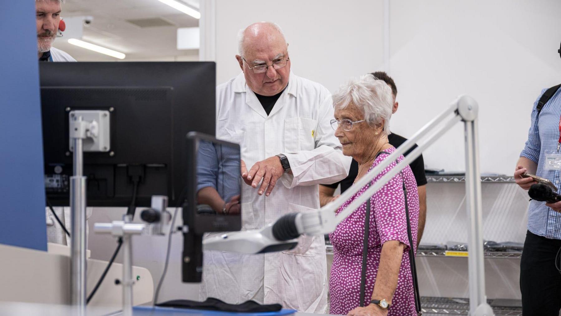 A middle aged man talks to an elderly woman wearing a pink dress while she looks at a screen in an engineering space.