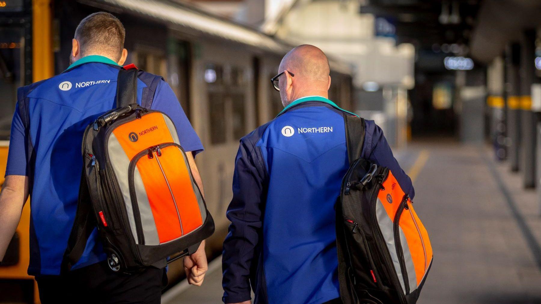 Two employees wearing blue Northern-branded uniforms walk with their backs to the camera. Both are also wearing orange and silver Northern-branded rucksacks.