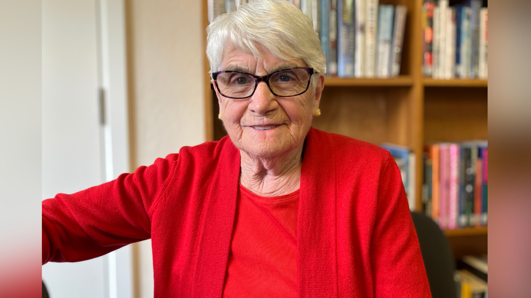 Patricia Dalton pictured at the coffee morning in Wye, near Ashford in Kent. Patricia is wearing a red jumper and black glasses. She has white hair and is smiling at the camera.
