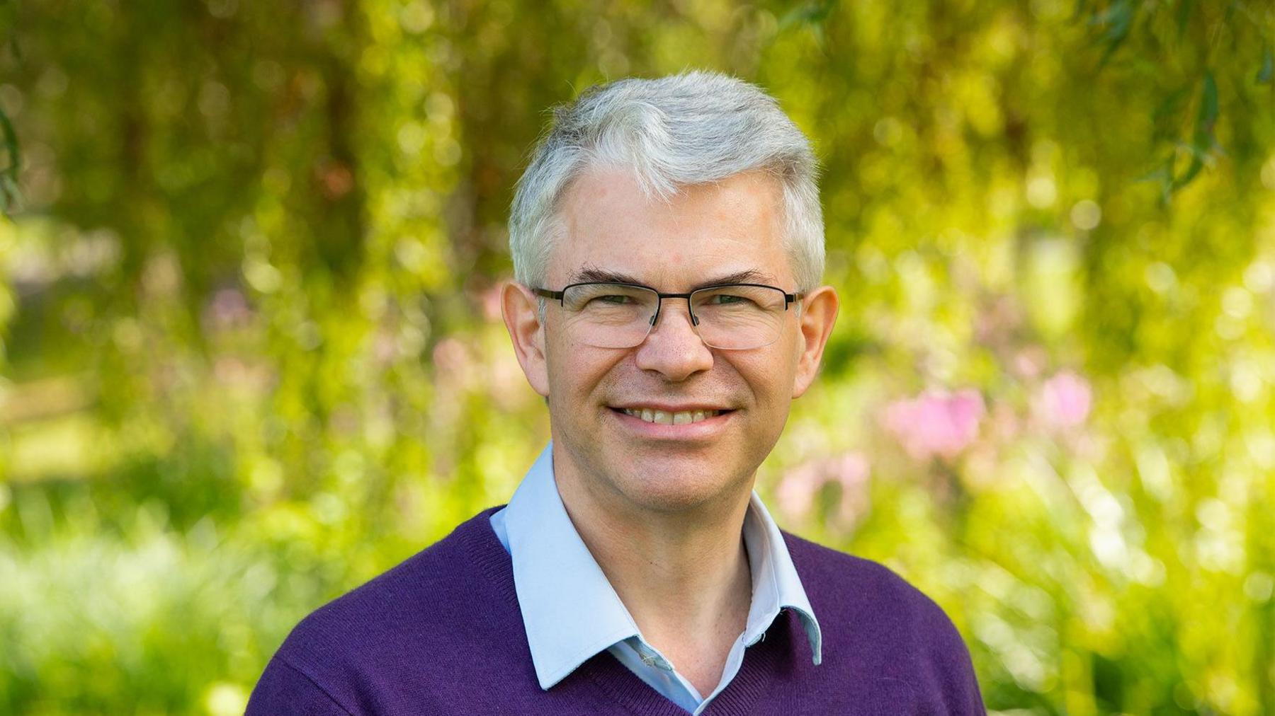 A man in a purple jumper with a blue shirt underneath, he is wearing glasses and has grey hair. The background is blurred but he is outside next to some green plants.