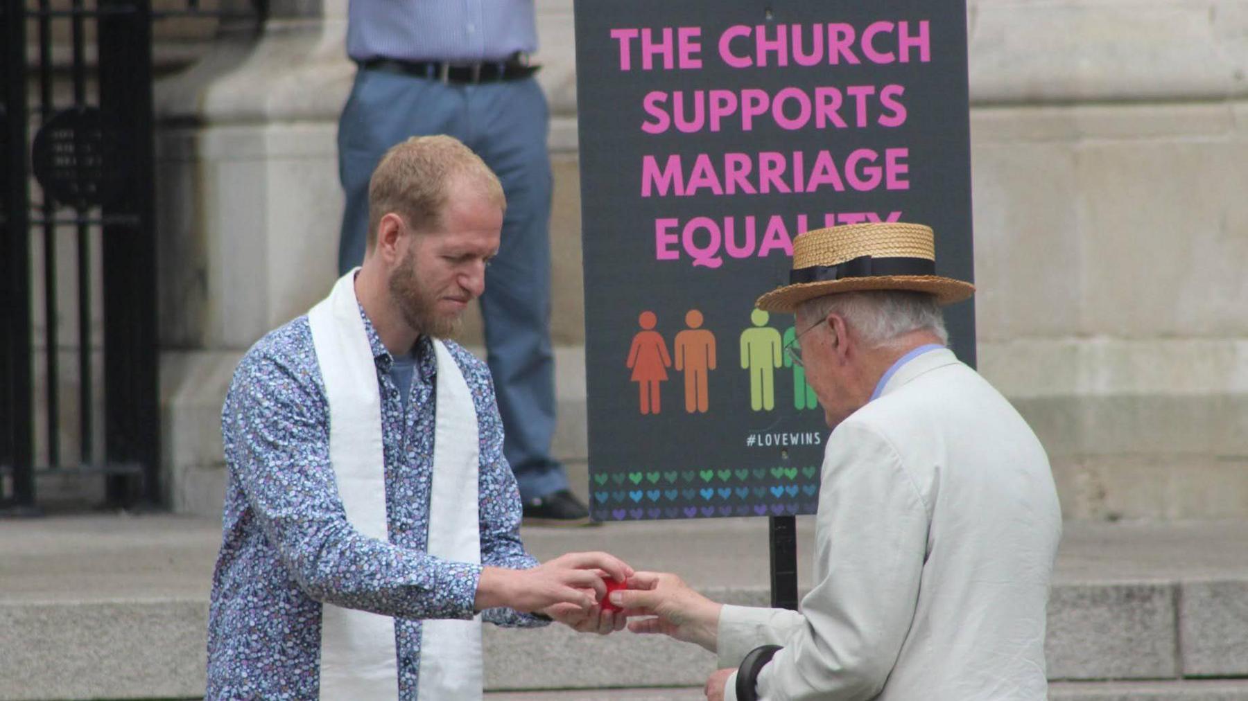 Pastor Steve Ames, who is wearing a blue and white paisley print button shirt with a white stole over his neck, hands an older man wearing a flat top hat and light coloured suit jacket a small red cup. They are stood outside a white stone building beside a sign that says 'The church supports marriage equality'.