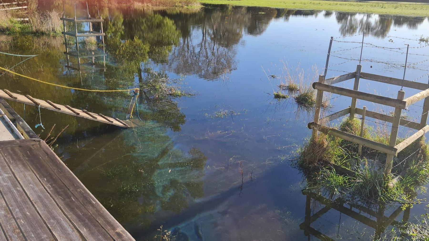 A field next to a moored boat completely submerged in water. A gangplank leads from the edge of the boat, with the other end underwater.