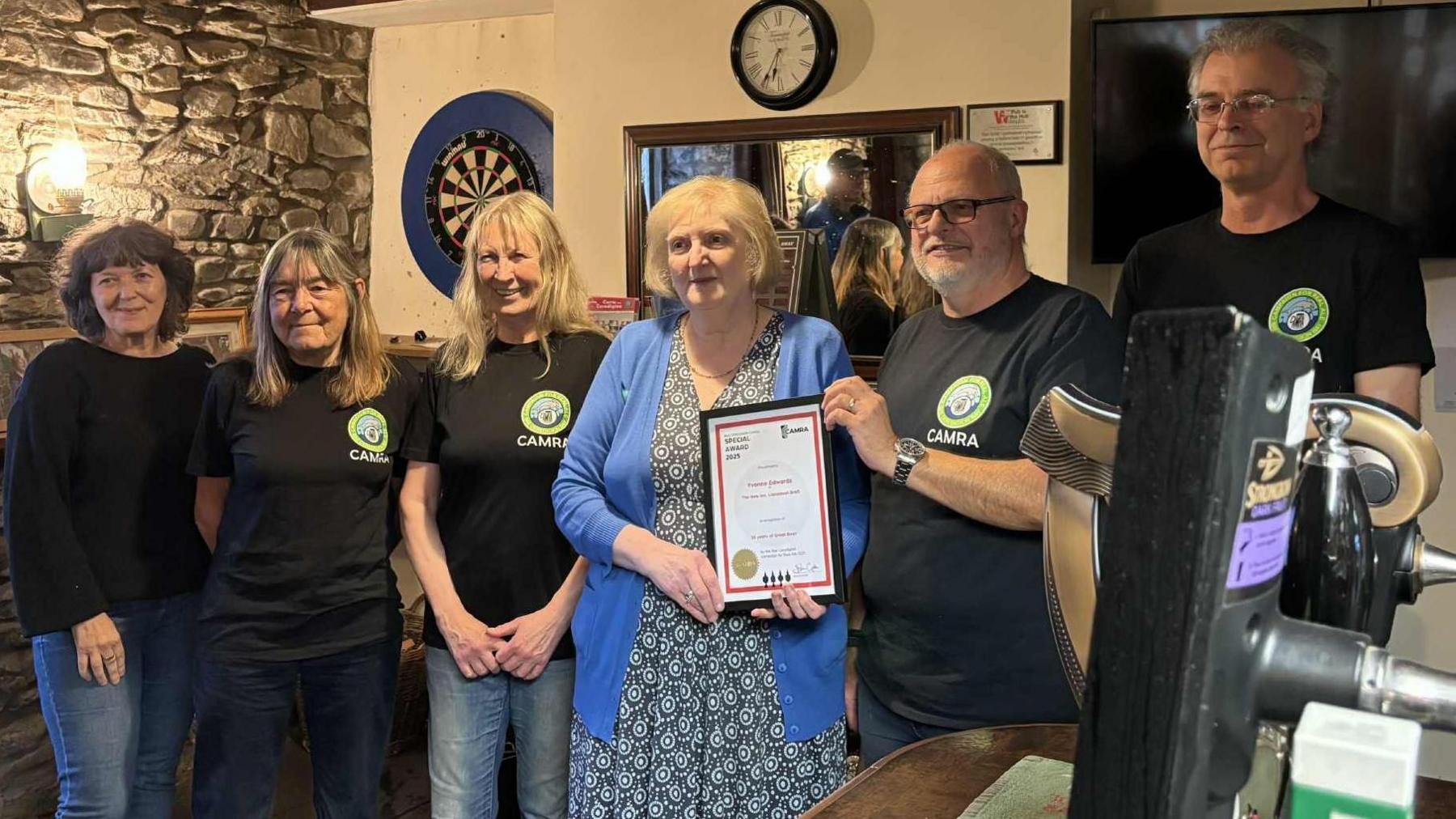 Llun o aelodau CAMRA gan gynnwys y Cadeirydd John Gale yn gwobrwyo tystysgrif i Yvonne. Mae'r chwech ohonynt yn sefyll ger y bar yn y New Inn, gyda golau cynnes a bwrdd darts yn y cefndir.