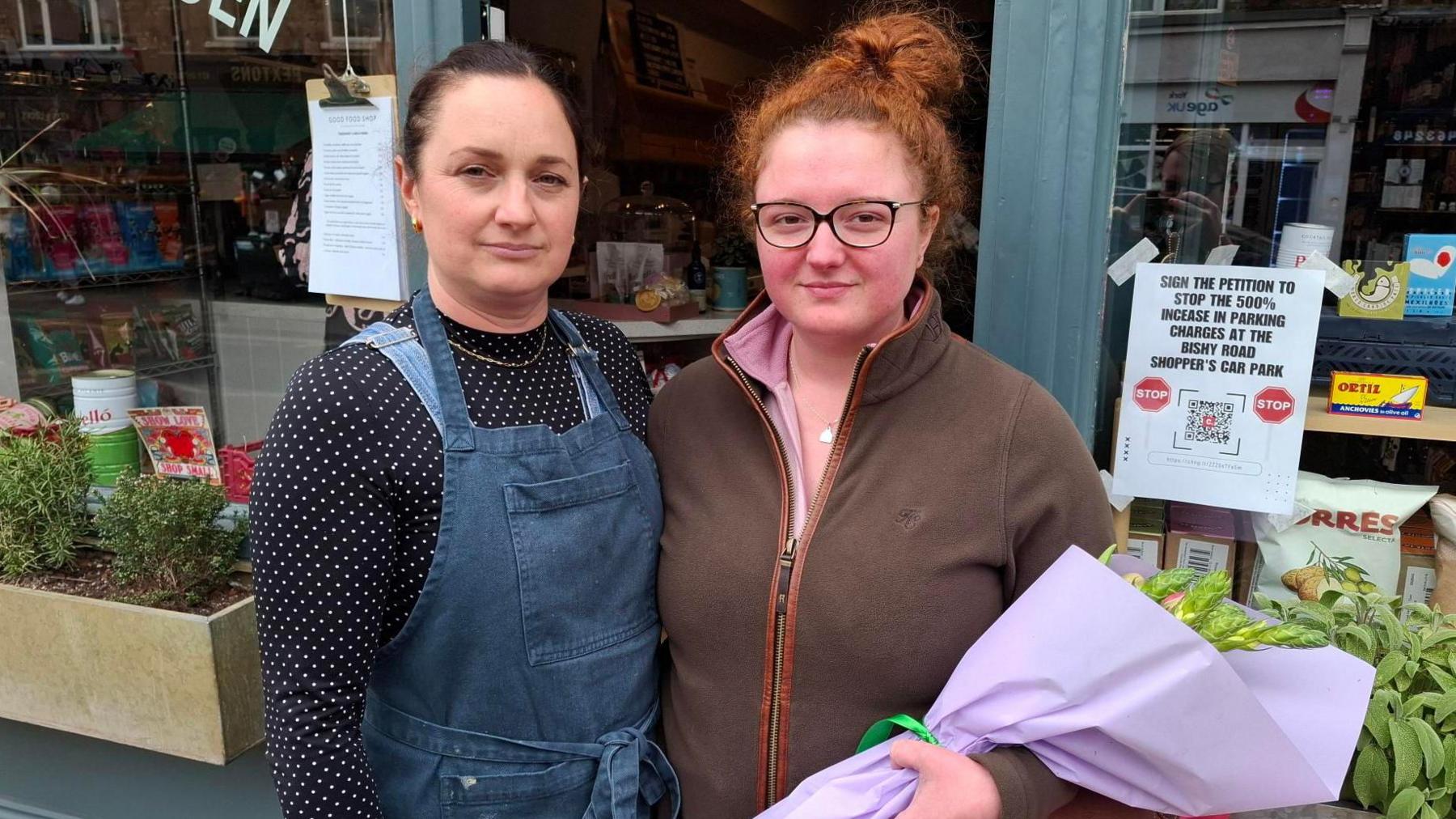Ruth Phillips and Charlotte Kneafsey stand outside a shop looking at the camera. Ruth is wearing a polka-dot T-shirt under a denim apron, while Charlotte is wearing a pink shirt under a brown fleece. Charlotte is also carrying a bouquet of flowers.