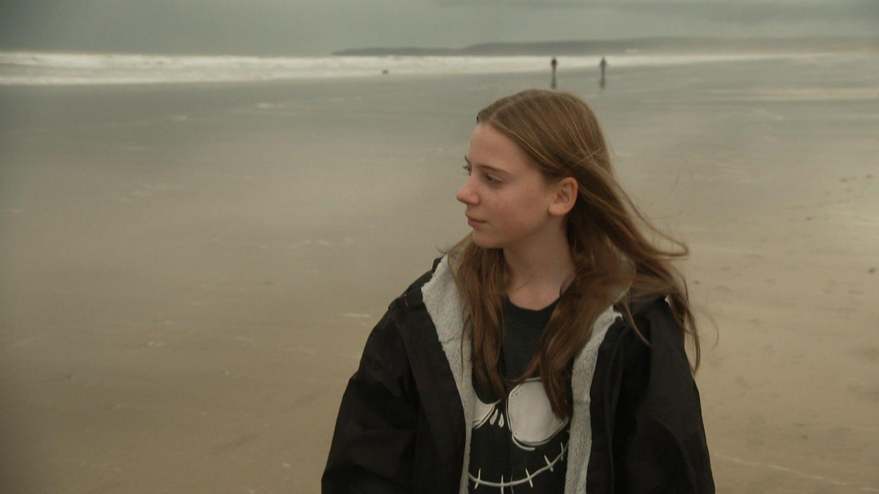 A young girl walking on the beach in a fur-lined black coat, with brown hair, looking out into the ocean.