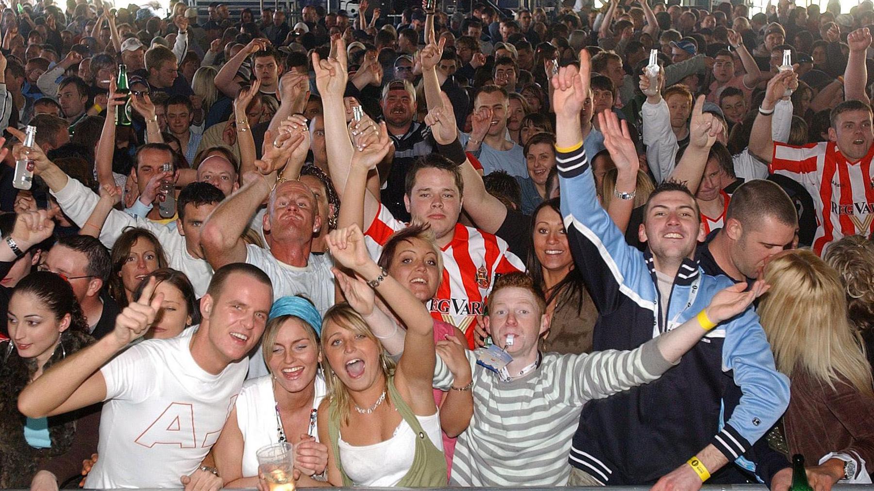 A picture taken from the stage of a crowd of people singing and dancing with arms in the air. Several are wearing red and white-striped Sunderland shirts, in one corner there is a couple kissing. A man at the front with short ginger hair is blowing a whistle, there are two young women with blonde hair cheering next to him and many people are clutching plastic drink glasses and bottles.