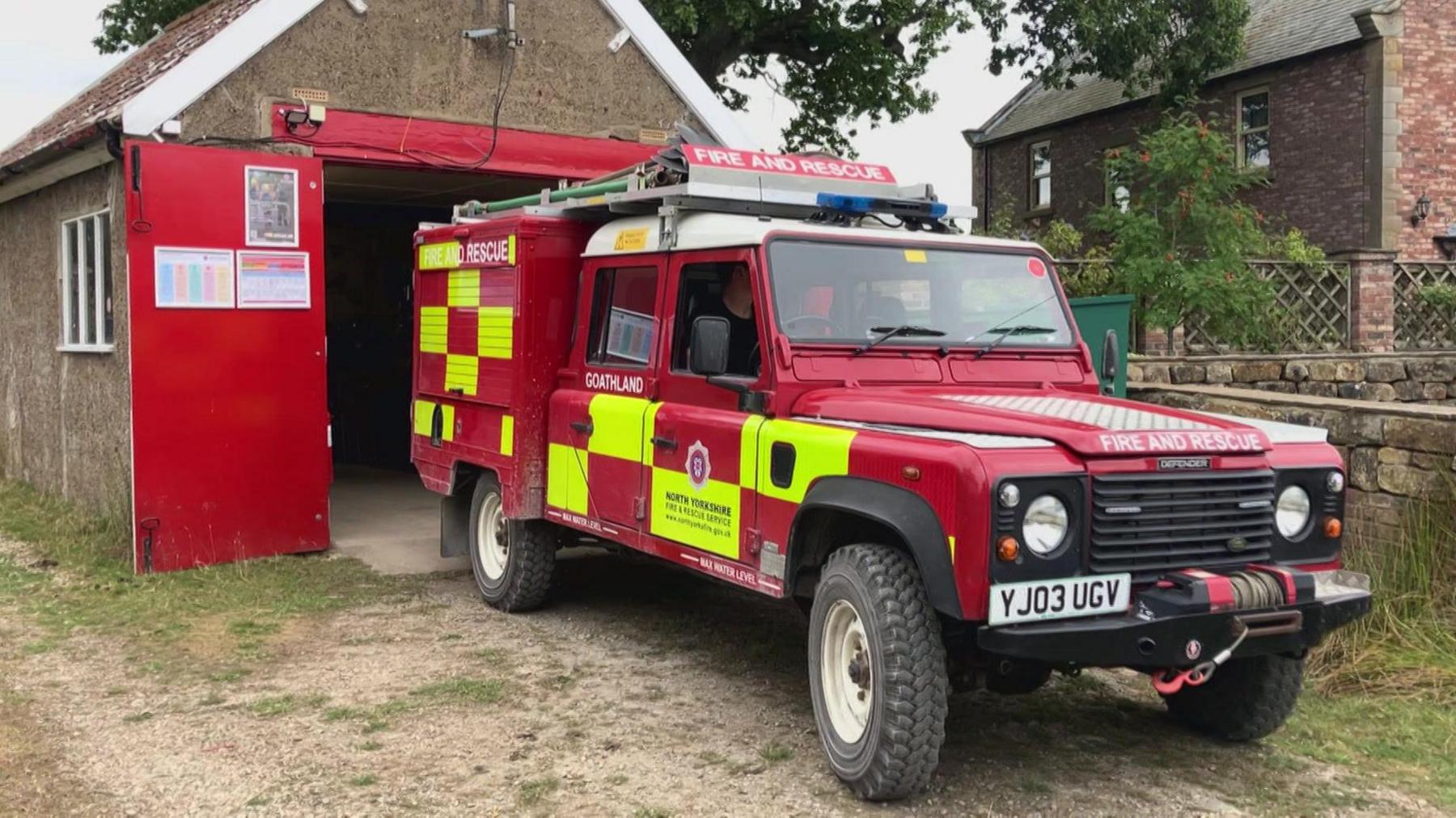 A red and yellow chequered Land Rover with blue lights on the top. It reads "Fire and Rescue" on the front and "Goathland" on the side. The back has cupboards secured by shutters. It is outside a shed with large red doors.