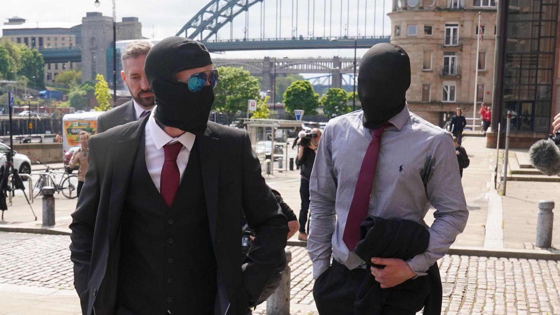 Graham and Carruthers walk into court. Both are wearing suits and have black hoods covering their faces