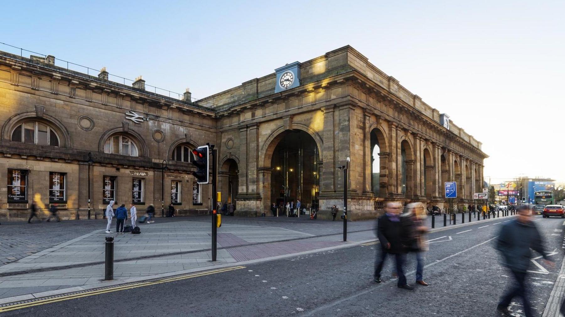 Photo of Newcastle Central Station with the sun about to go down. It is a grand brick building with several arches and a clock on the top. There are people crossing the road and cars on the road. There are people standing outside the station.