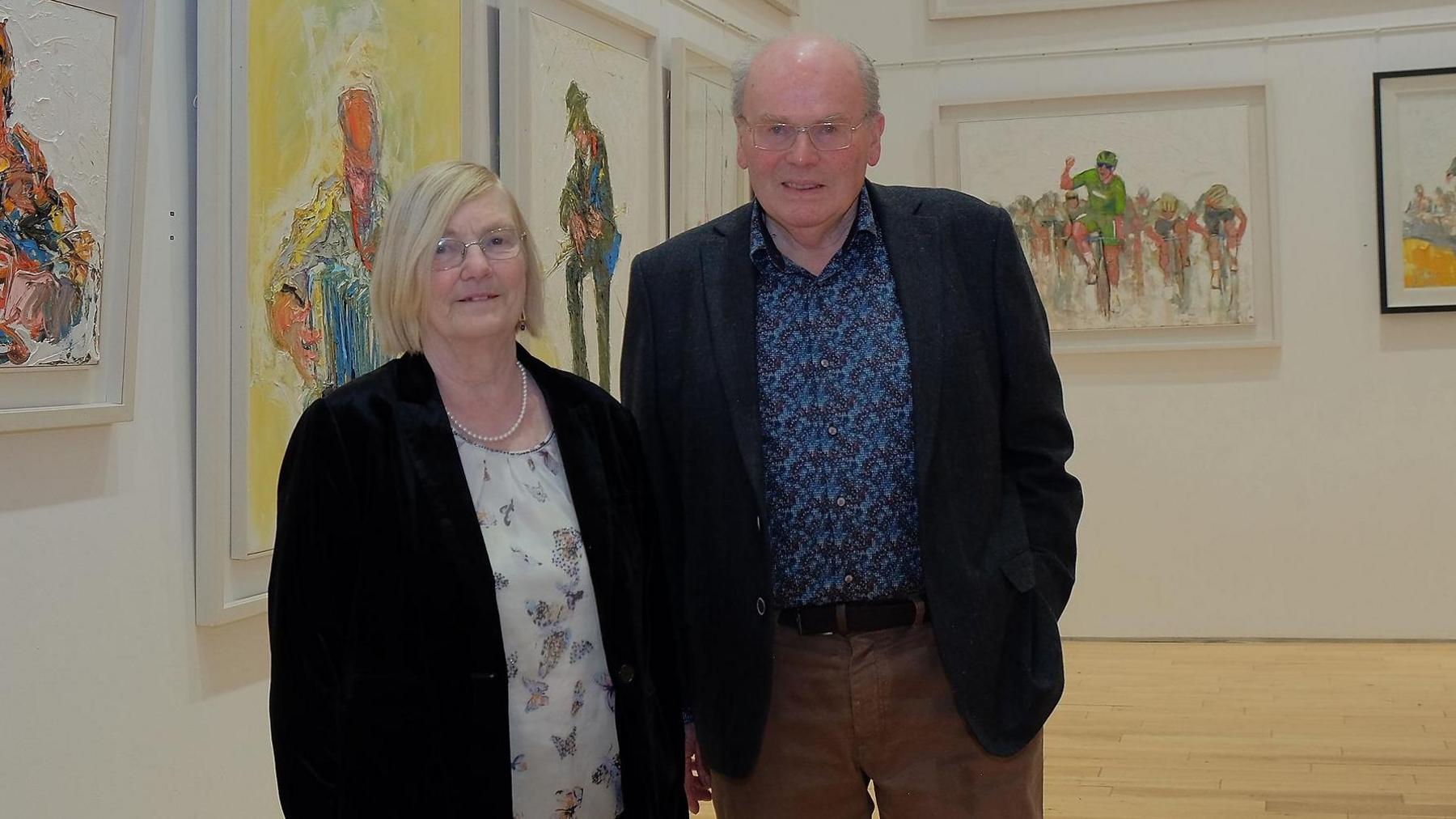 A man and woman are standing in an art gallery. They are both wearing glasses and smiling at the camera.
