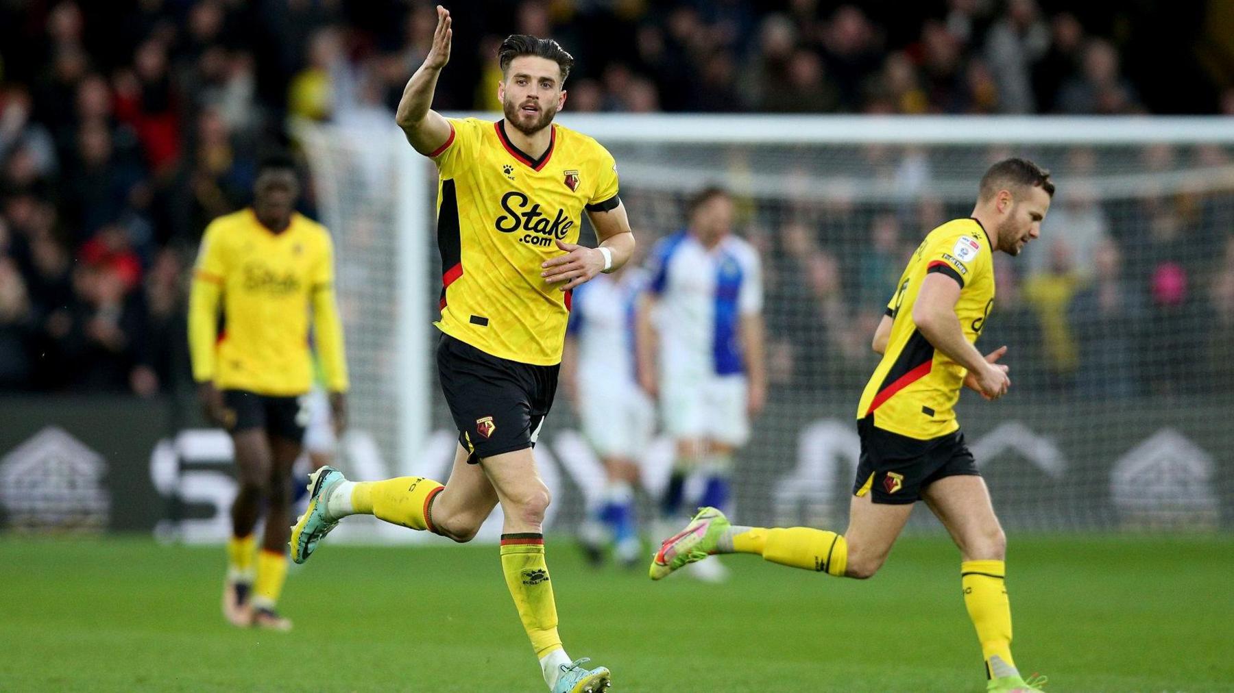 Watford's Wesley Hoedt celebrates after scoring his side's first goal during a Championship match at Vicarage Road, Watford, in February 2023.
He wears the Watford home kit - bright yellow - and holds a hand aloft as he runs across the pitch, with another team-mate nearby. Their opponents are blurry in the background, in blue and white. 