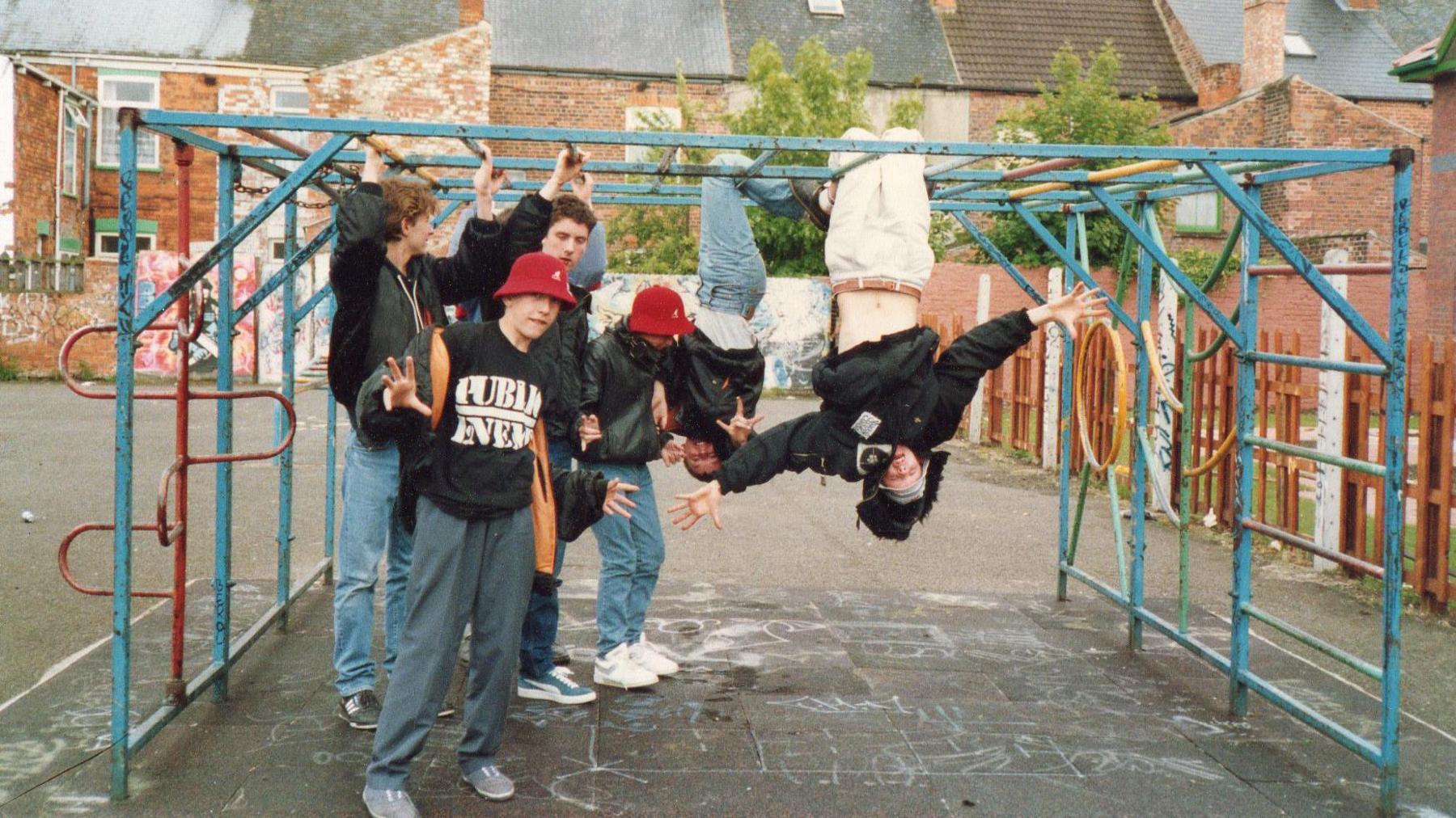 Six boys playing on a metal climbing frame, which is on tarmac with a row of terraced houses in the distance