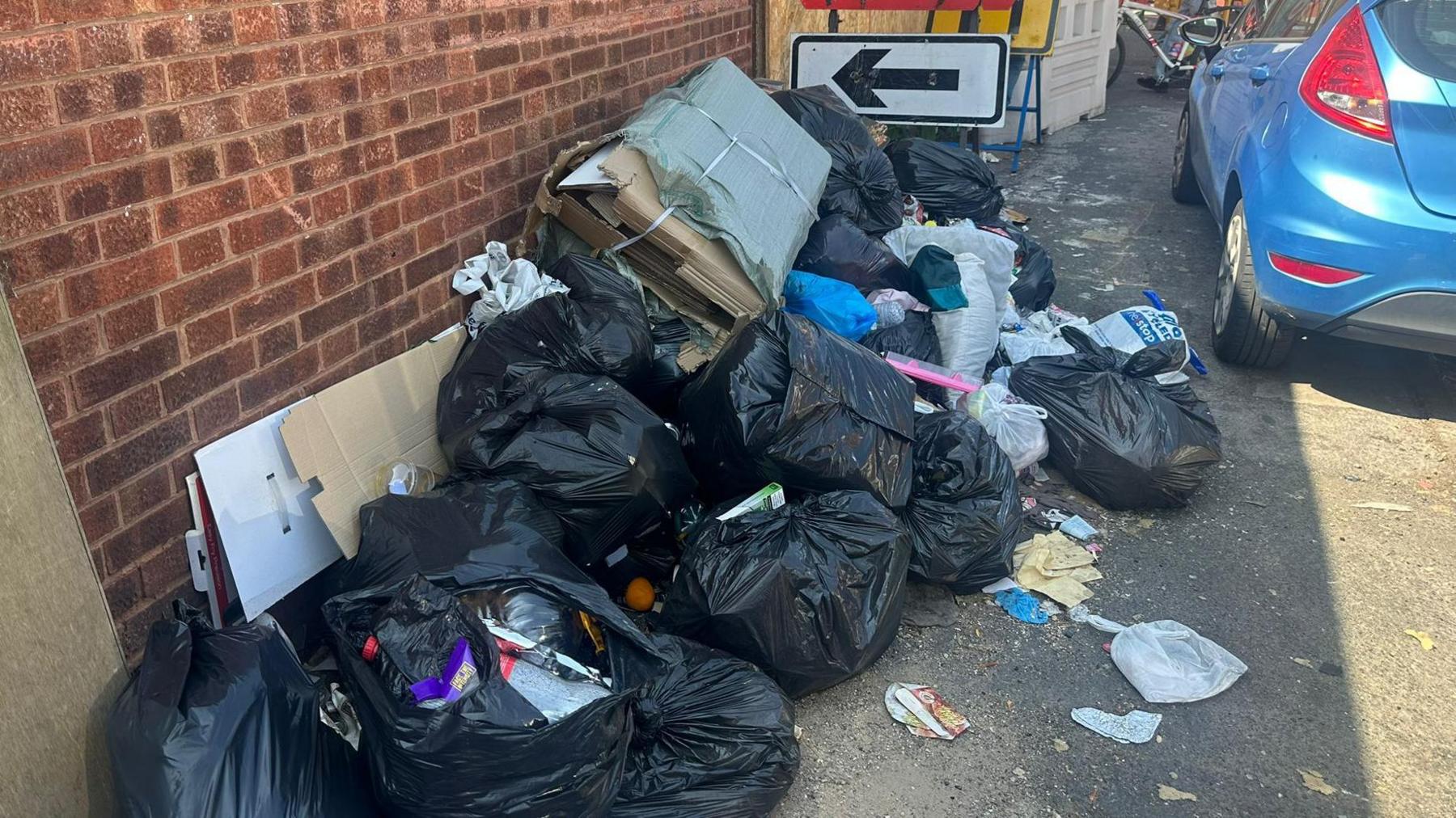 A pile of rubbish on a pavement in Birmingham, with roadwork signs in the background. The rubbish is almost completely blocking the pavement. There is a blue car parked alongside it.