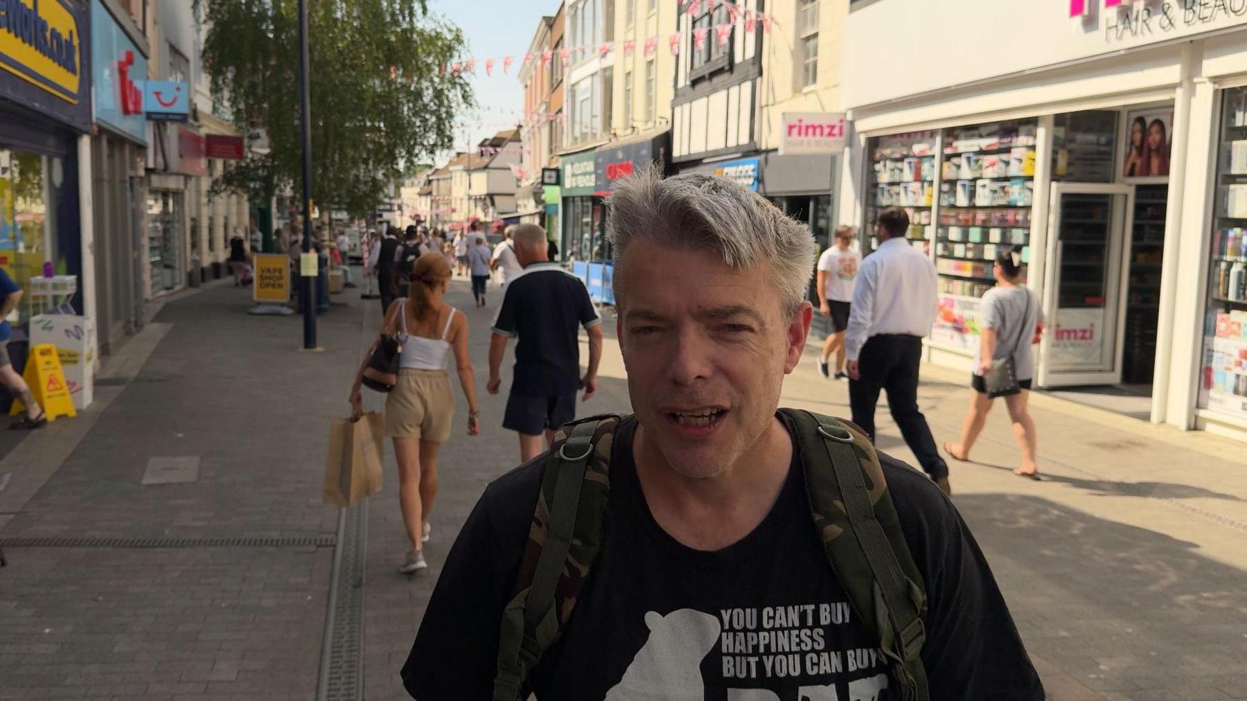 A picture of Danny Macauley in Maidstone town centre. He has a camouflage backpack, and is wearing a black t-shirt. He has greying hair. A number of shops, including The Works and Greggs can be seen in the background.