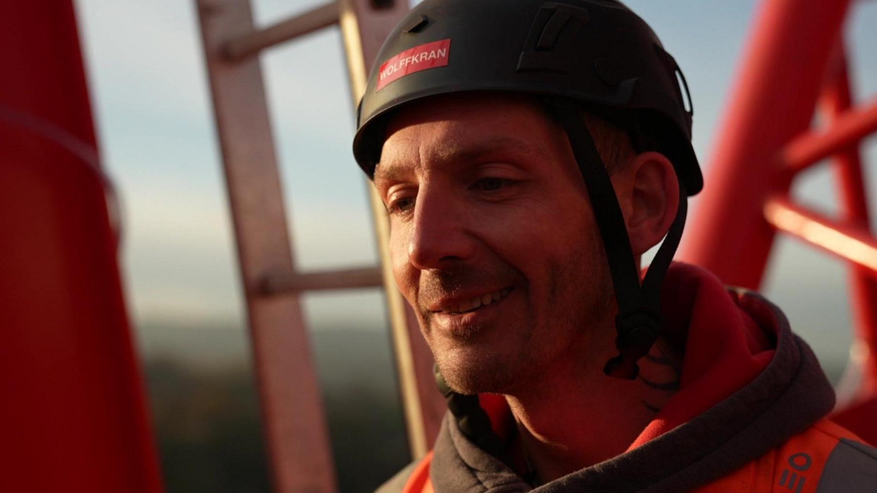 A man in a black hard hat stands on a crane wearing black and orange safety clothing.