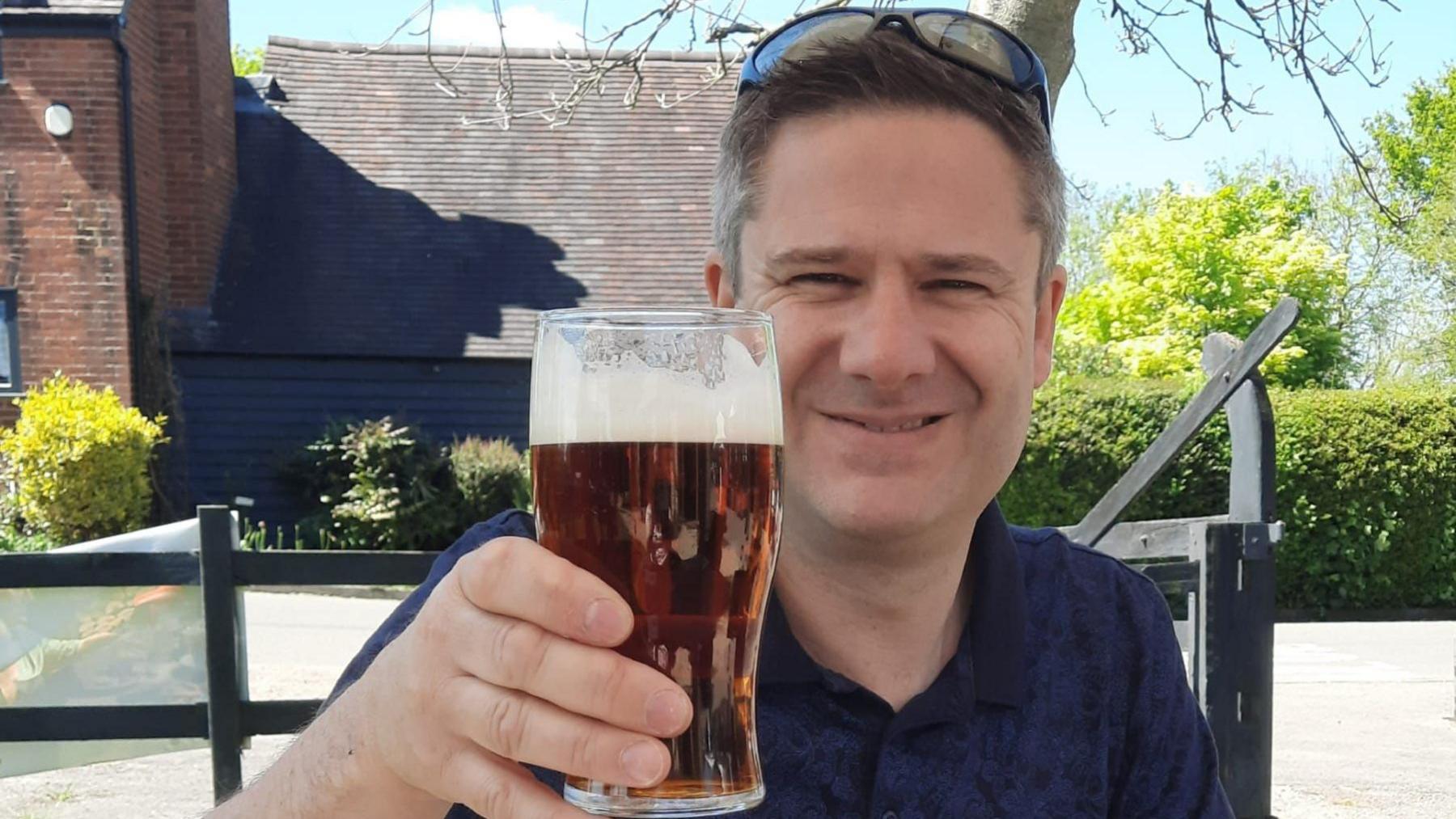 Andy Probert sitting in a pub garden while holding a pint of beer up in his right hand. He is smiling while wearing a navy polo shirt and has sunglasses on top of his head. Behind him is a black weatherboarded building on a sunny day.