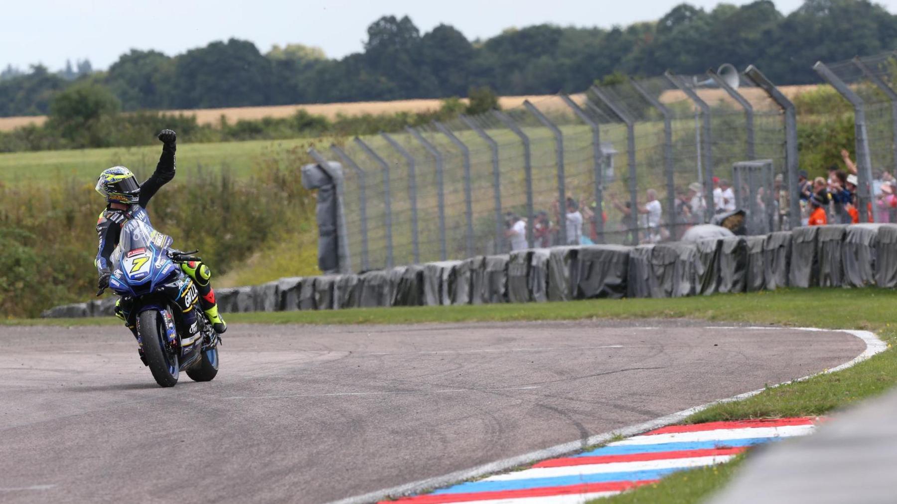 Ryan Vickers riding on his bike in multi-coloured OMG Racing Yamaha gear and helmet. His arm is in the air to acknowledge a crowd after a race at the British Superbikes.