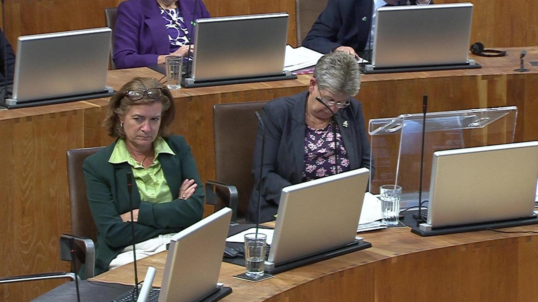 Eluned Morgan sitting in the Senedd chamber with her arms folded next to Jane Hutt, who has her head down